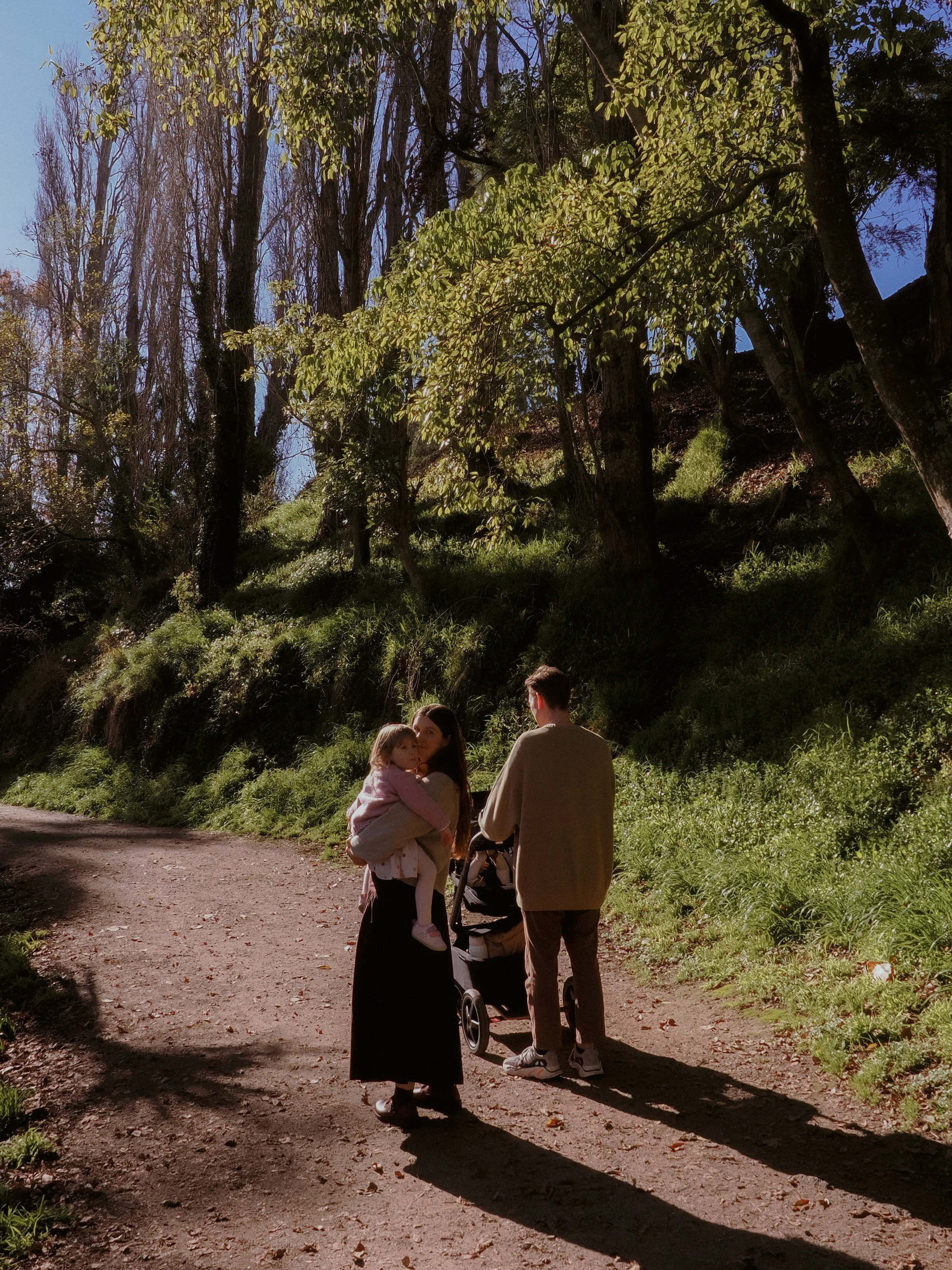 A family of three, including a woman holding a young girl, a man with a stroller, and a child in a stroller, walking on a dirt path in a wooded area during daytime with sunlight filtering through trees.