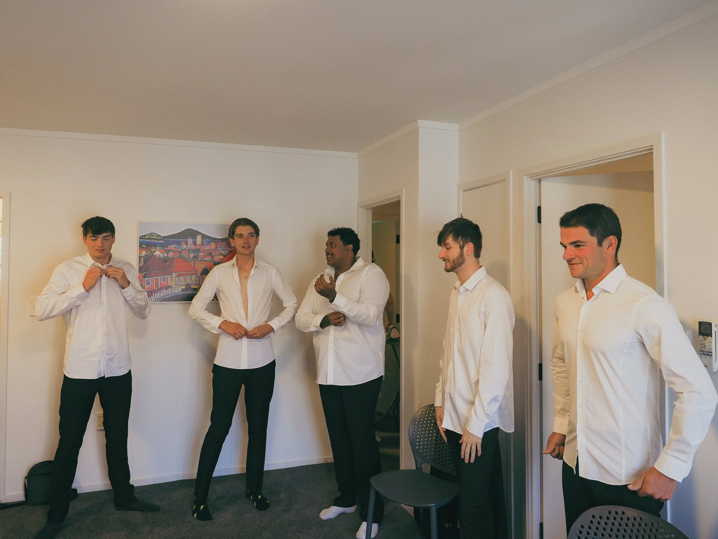 Five young men in white shirts standing in a room, engaging in conversation and adjusting their shirts, with a painting of a cityscape and mountains on the wall behind them.