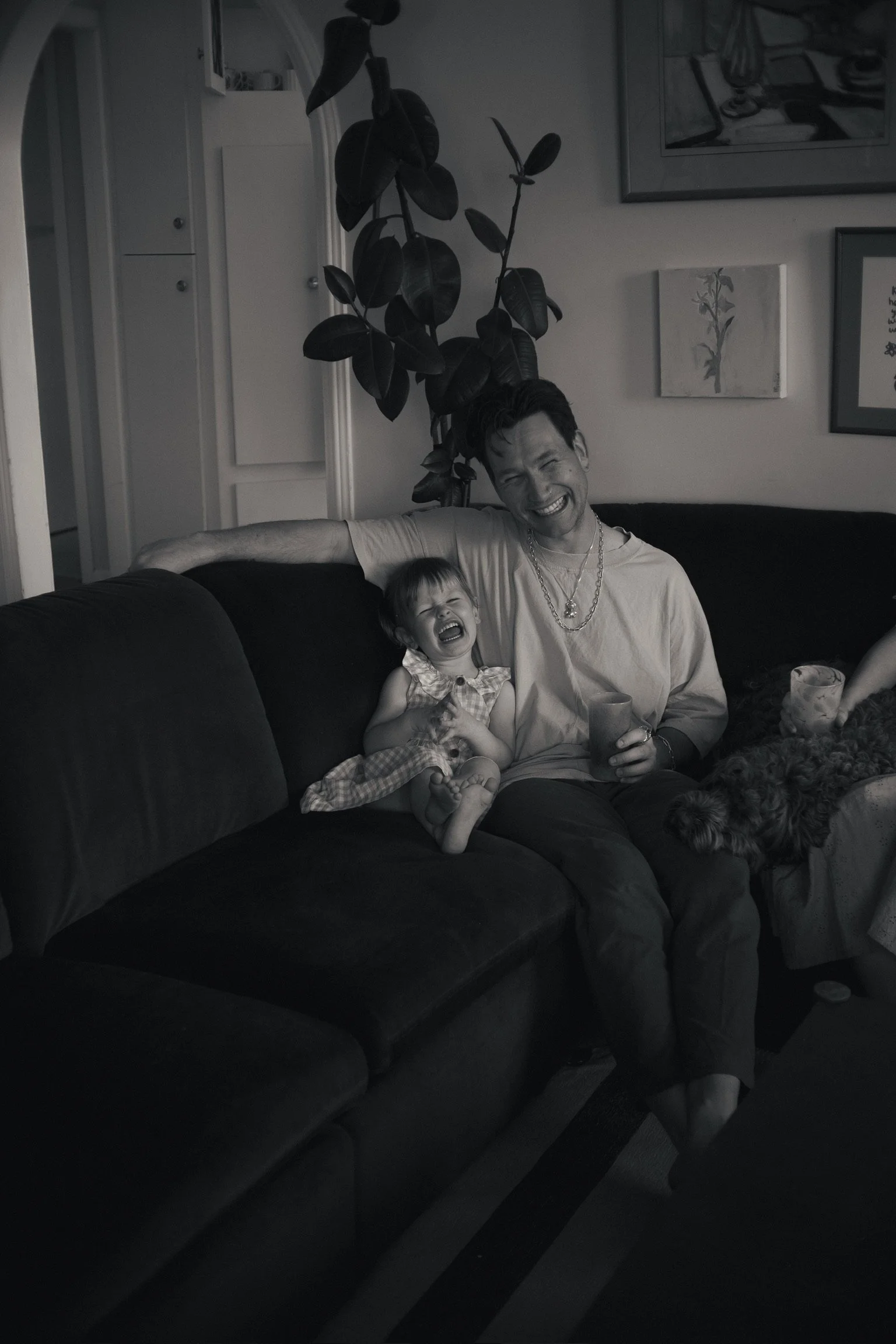 A man and a young girl sitting on a couch, both laughing and appearing happy, with a dog lying beside them. The room has framed pictures on the wall and a large potted plant in the background. Family Photographer. Hamilton New Zealand