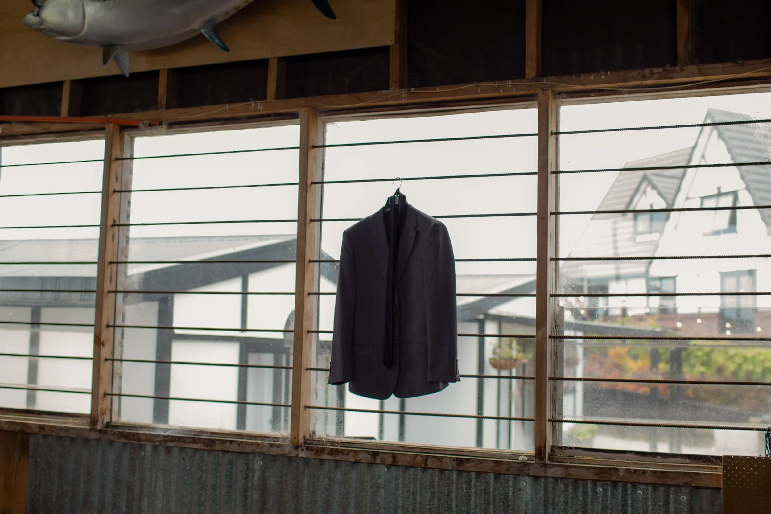 A suit jacket and tie hanging in front of a window with horizontal bars, with a house visible outside.  Wedding Photography Waikato New Zealand. Wedding Photography Hamilton New Zealand. Northland Wedding Photography