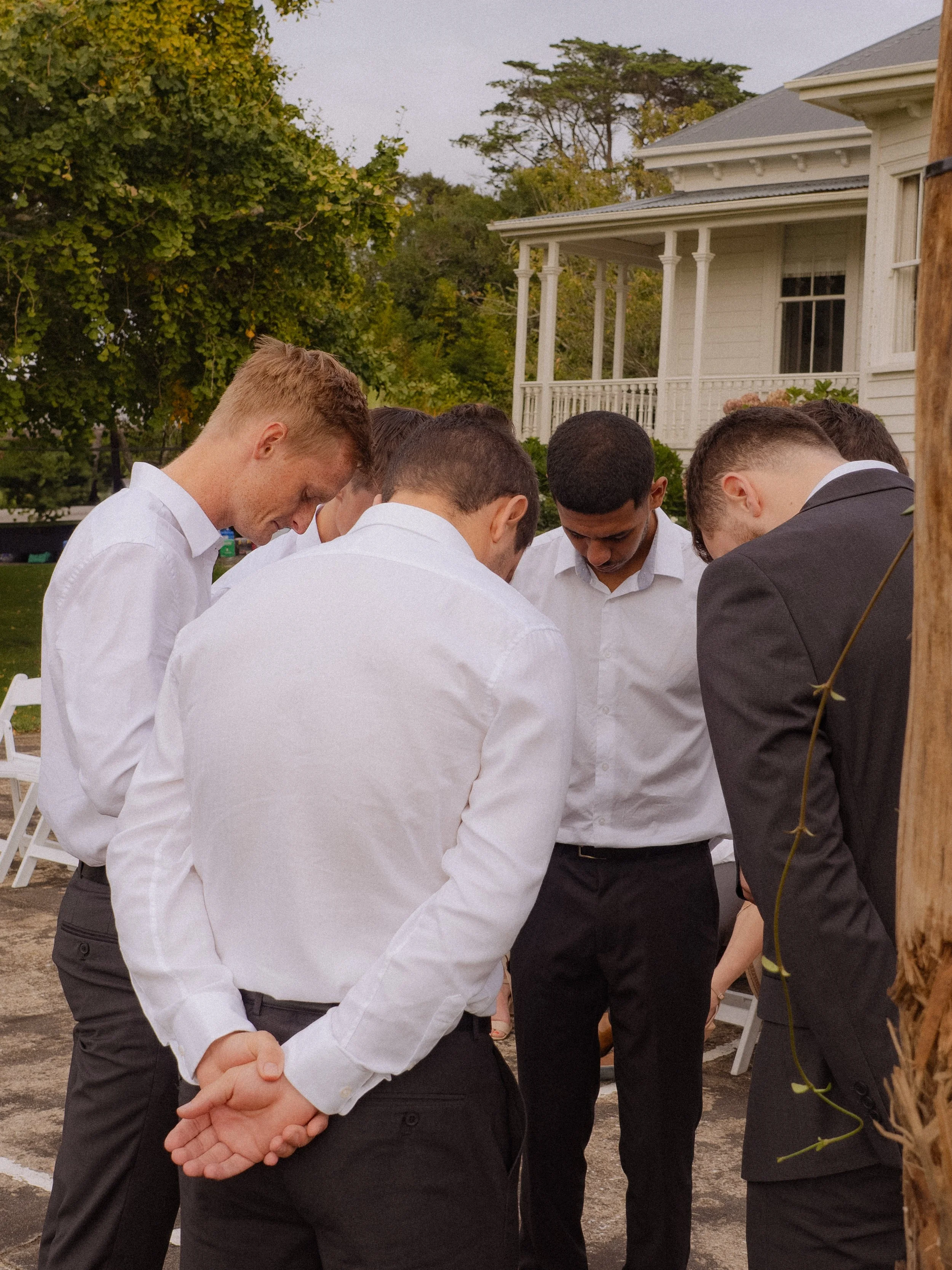 Group of young men dressed in formal attire, standing outdoors in a circle, with heads bowed and hands clasped, likely engaging in a prayer or moment of reflection.  Wedding Photography Waikato New Zealand. Wedding Photography Hamilton New Zealand. 