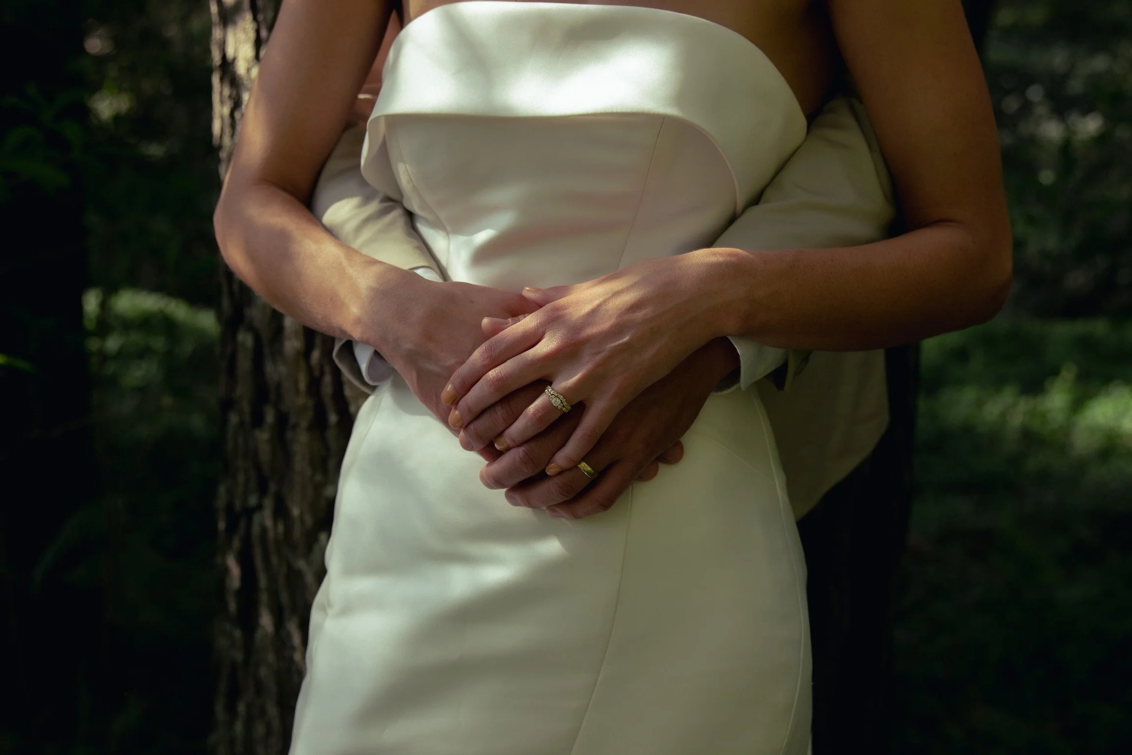 Close-up of a couple embracing outdoors; the woman is in a white dress with a ring on her finger, and the man's arm is around her.