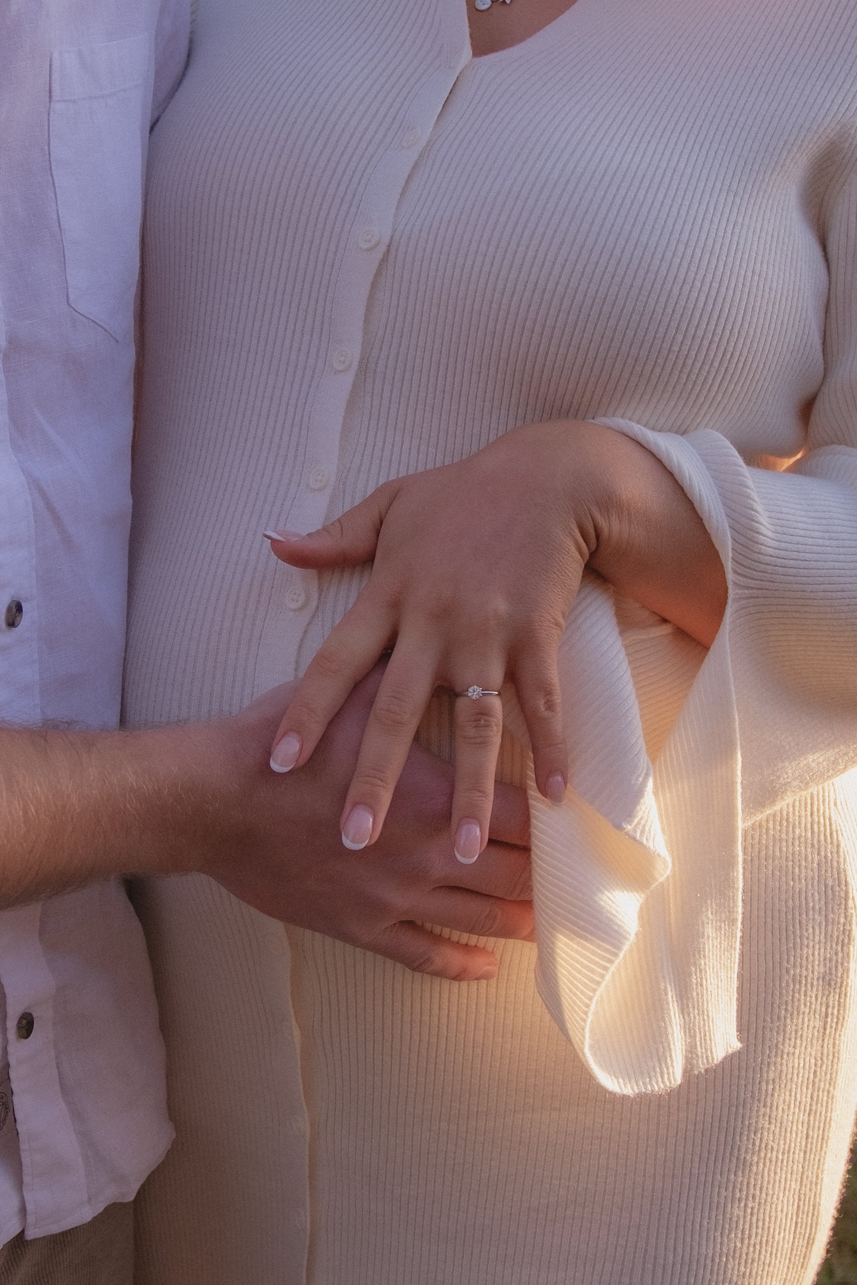 A close-up of a couple holding hands, with the woman wearing a ring on her finger showcasing her engagement or wedding ring. The woman is wearing a cream-colored, ribbed long-sleeve top, and the man is in a white, long-sleeved button-up shirt. Raglan