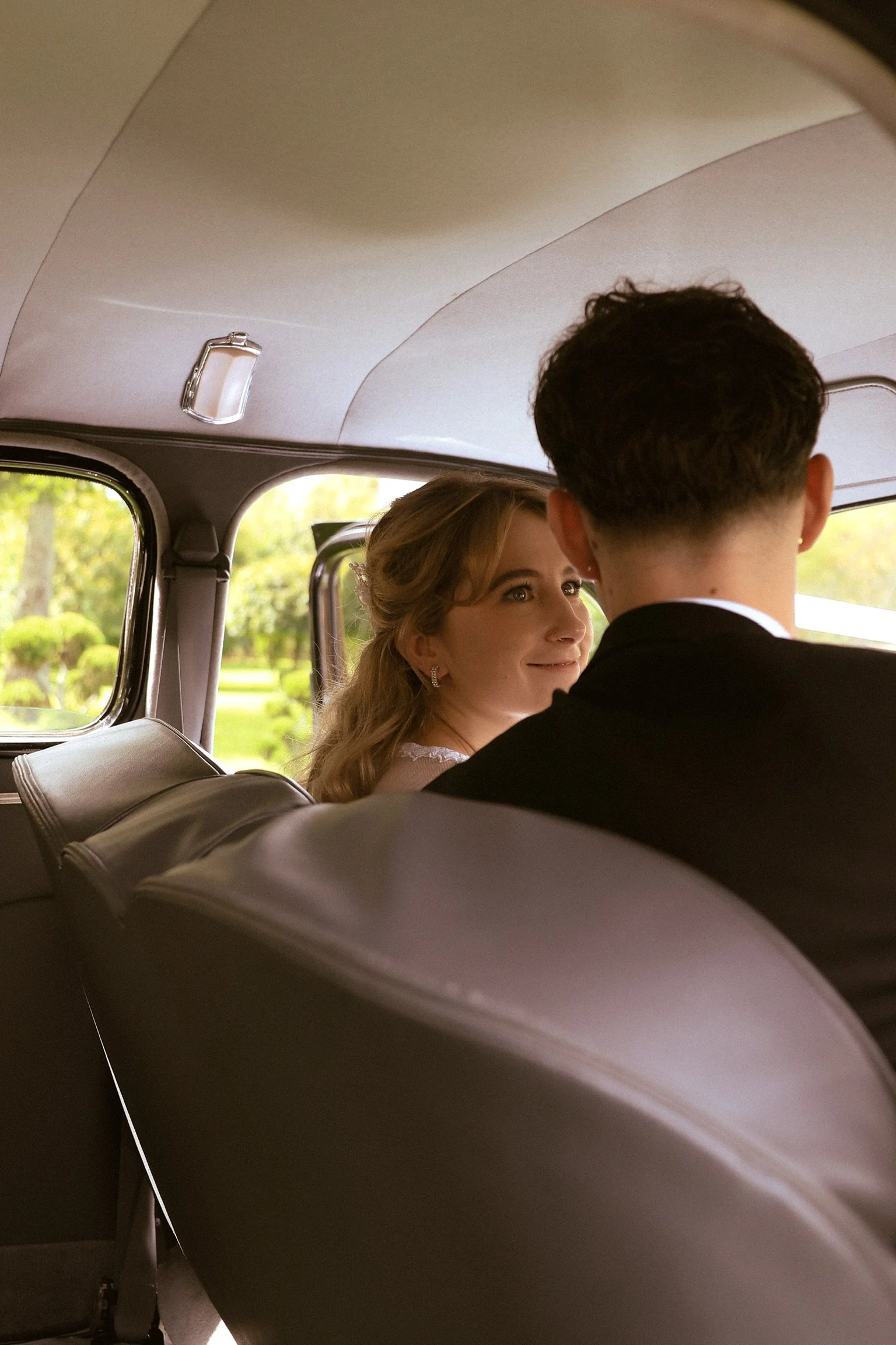 A bride and groom sit in the back of a vintage car, with the bride looking at the groom and smiling, amidst a lush green park scene outside the window. Wedding Photography Waikato New Zealand. Wedding Photography Hamilton New Zealand