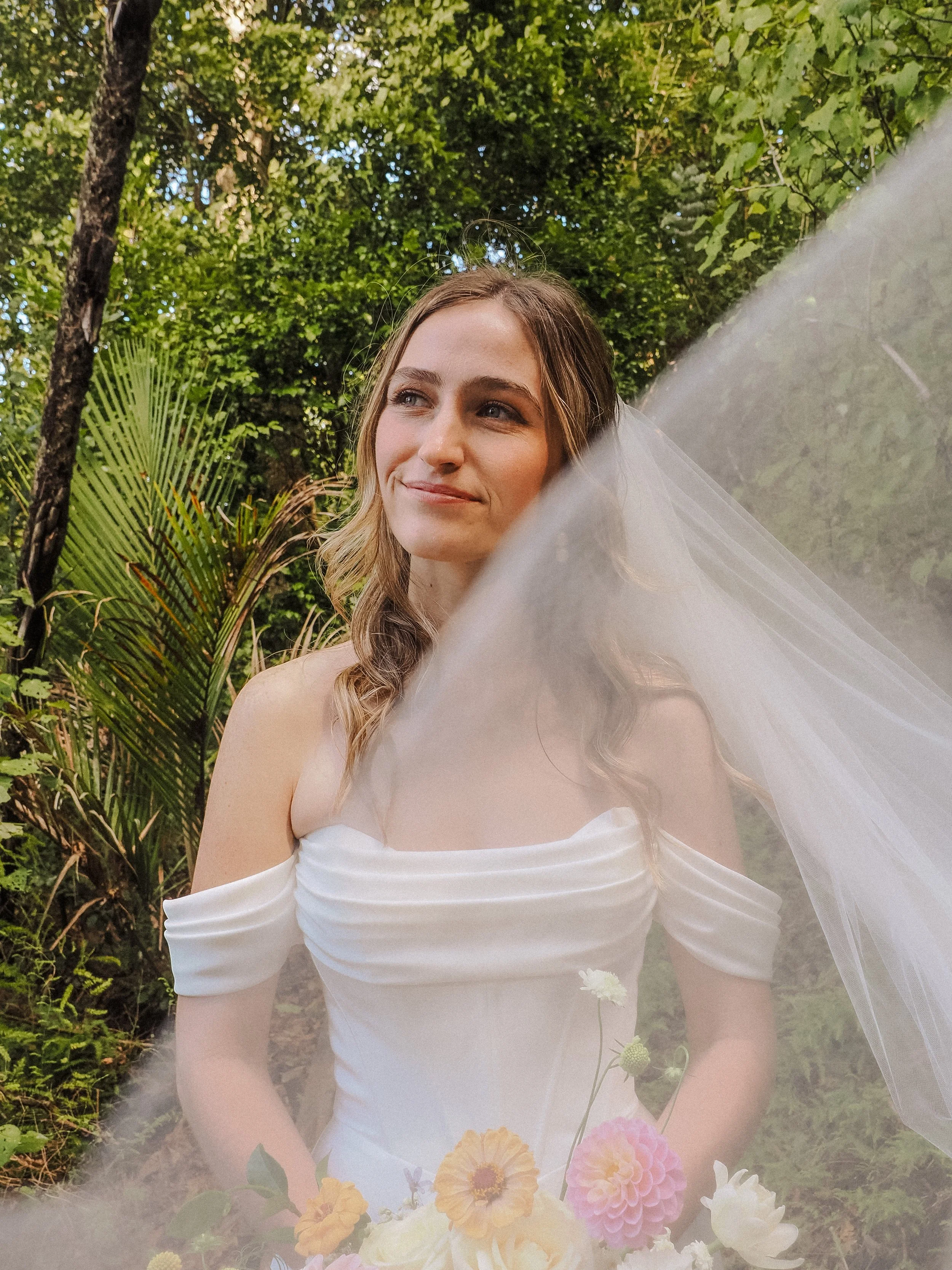 A bride with long wavy hair and a white off-shoulder wedding dress stands outdoors in a lush green setting, holding a bouquet of colorful flowers and gazing softly to her left, partially covered by a sheer veil.  Wedding Photography Waikato 