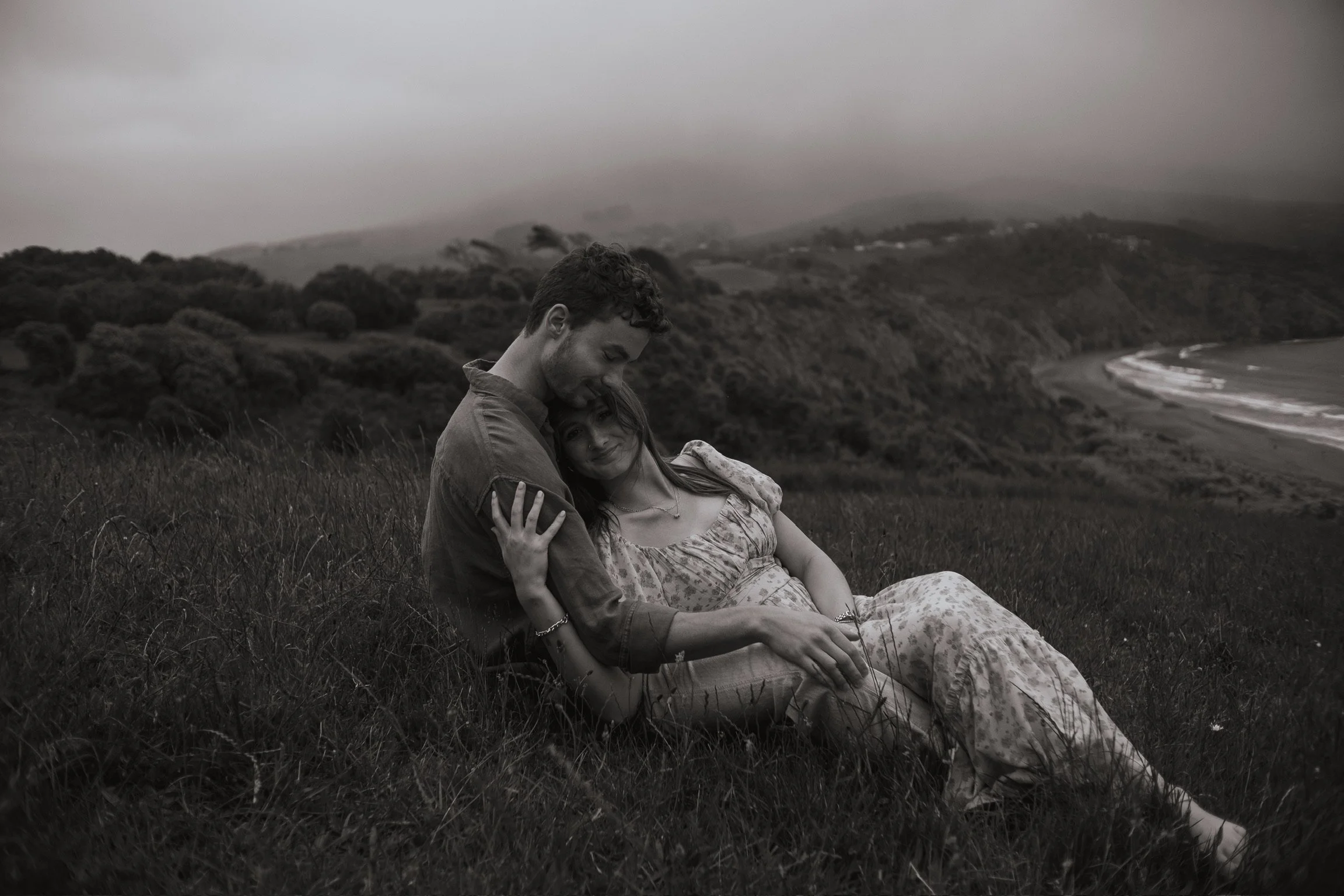 A black and white photograph of a couple sitting on a grassy hill near the coast, embracing, with an overcast sky and distant hills and shoreline in the background. Couples photography Raglan New Zealand. Engagement photography Hamilton New Zealand. 