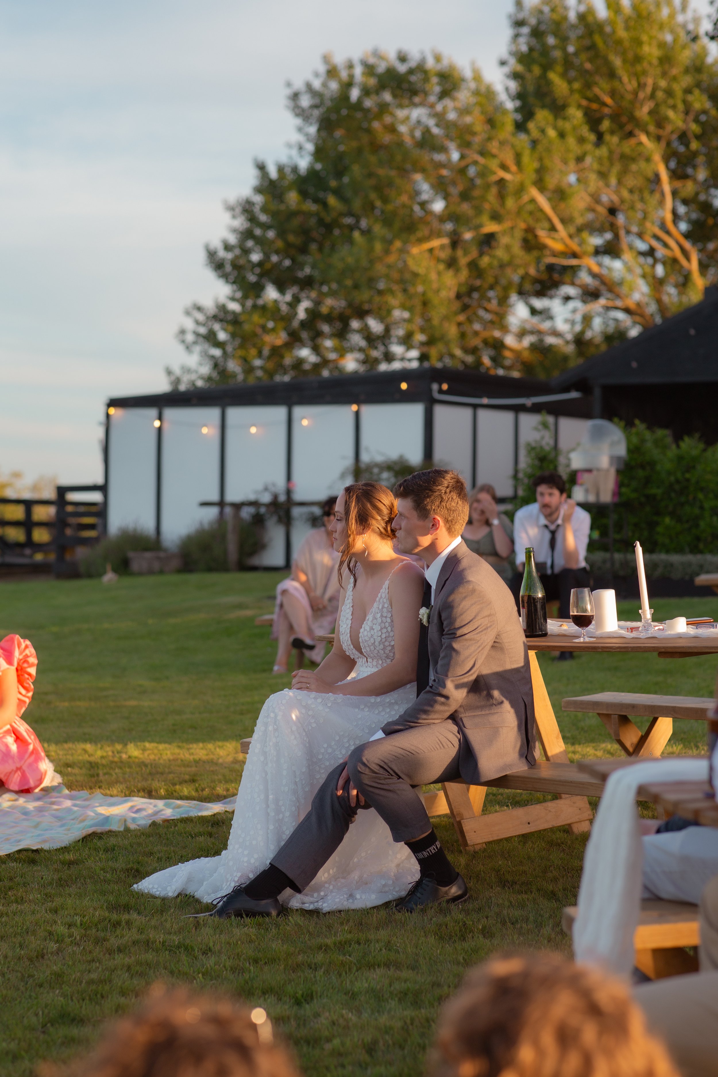 A bride and groom sitting on a wooden bench at an outdoor wedding reception during sunset, with guests and tables nearby and trees in the background.  Wedding Photography Waikato New Zealand. Wedding Photography Hamilton New Zealand. 