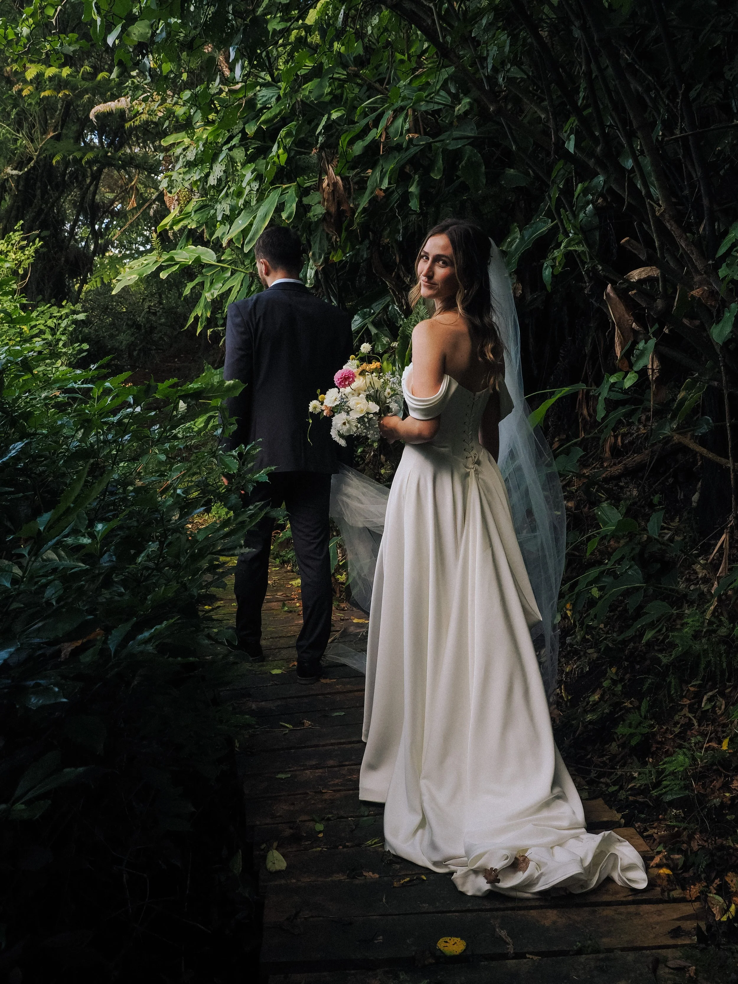 Bride in a white wedding dress holding a bouquet of flowers, standing on a wooden pathway in a lush, green forest with a groom in a dark suit facing away.  Wedding Photography Waikato New Zealand. Wedding Photography Hamilton New Zealand. Northland 