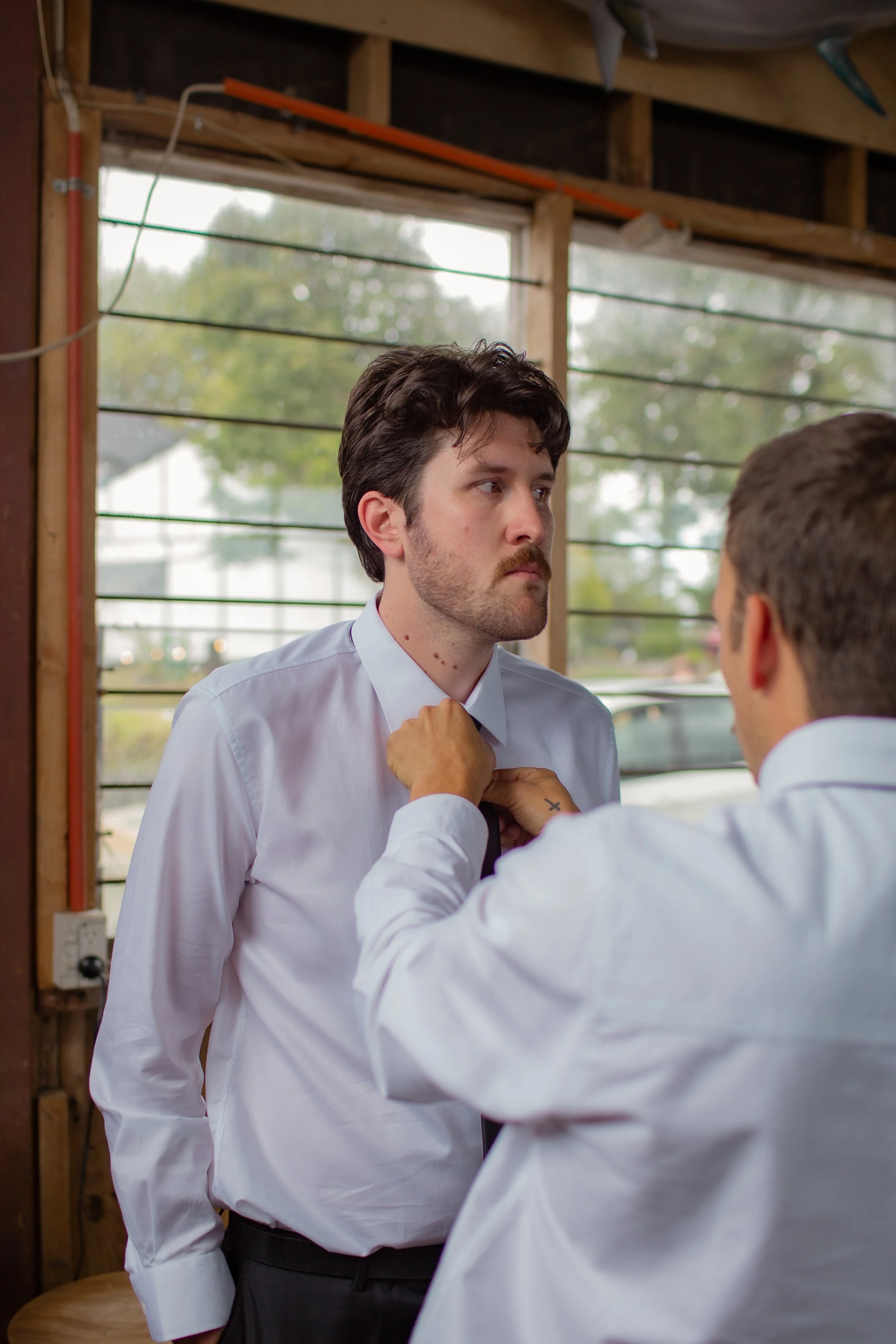 A man with dark hair and a beard wearing a white dress shirt stands still while another man adjusts his tie in what looks like a workshop or industrial space with large windows.