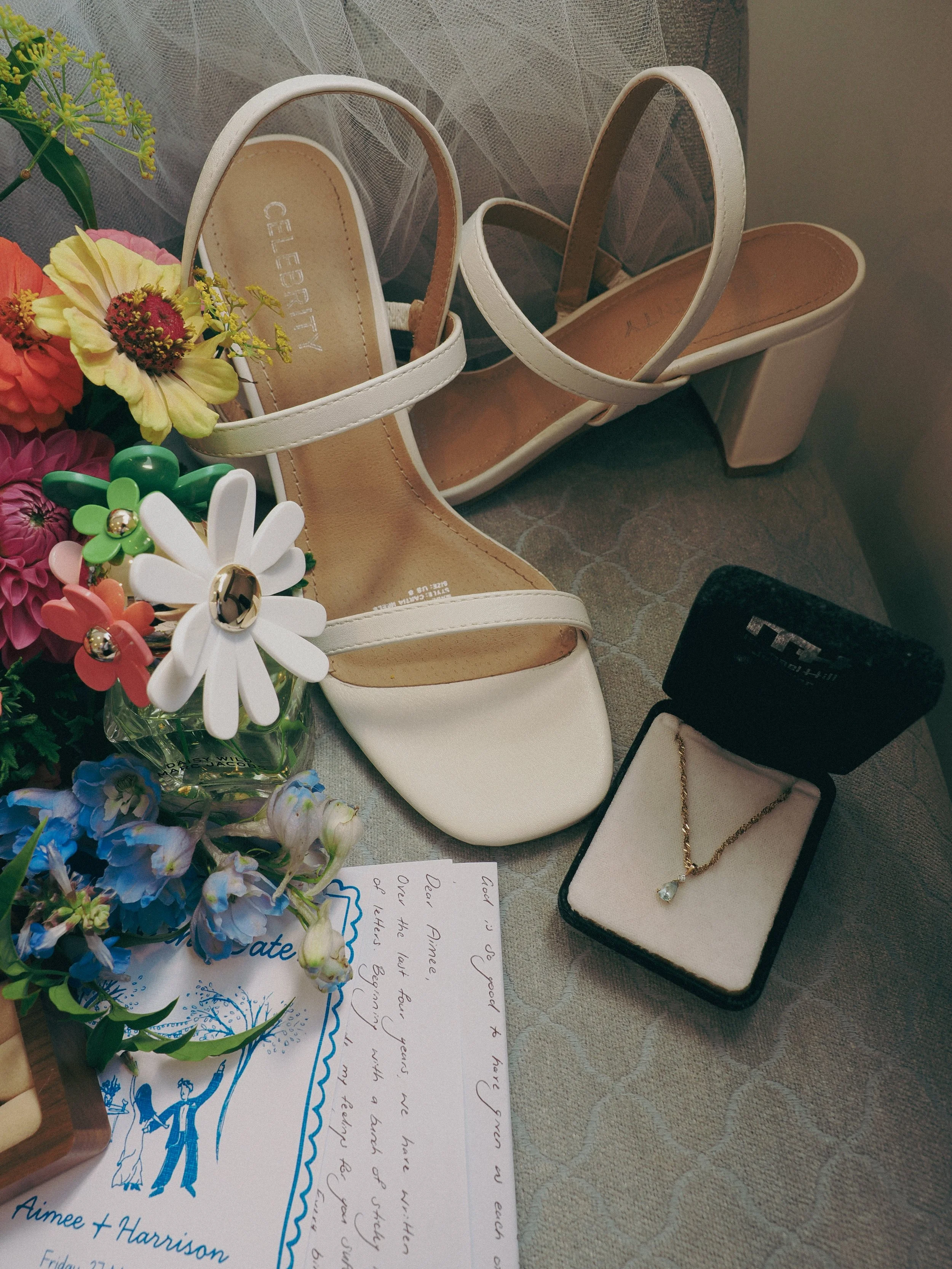 A pair of white high-heeled sandals with straps, a bouquet of colorful artificial flowers, a handwritten note, an open jewelry box with a gold necklace featuring a teardrop-shaped pendant, and a partially visible funeral program.