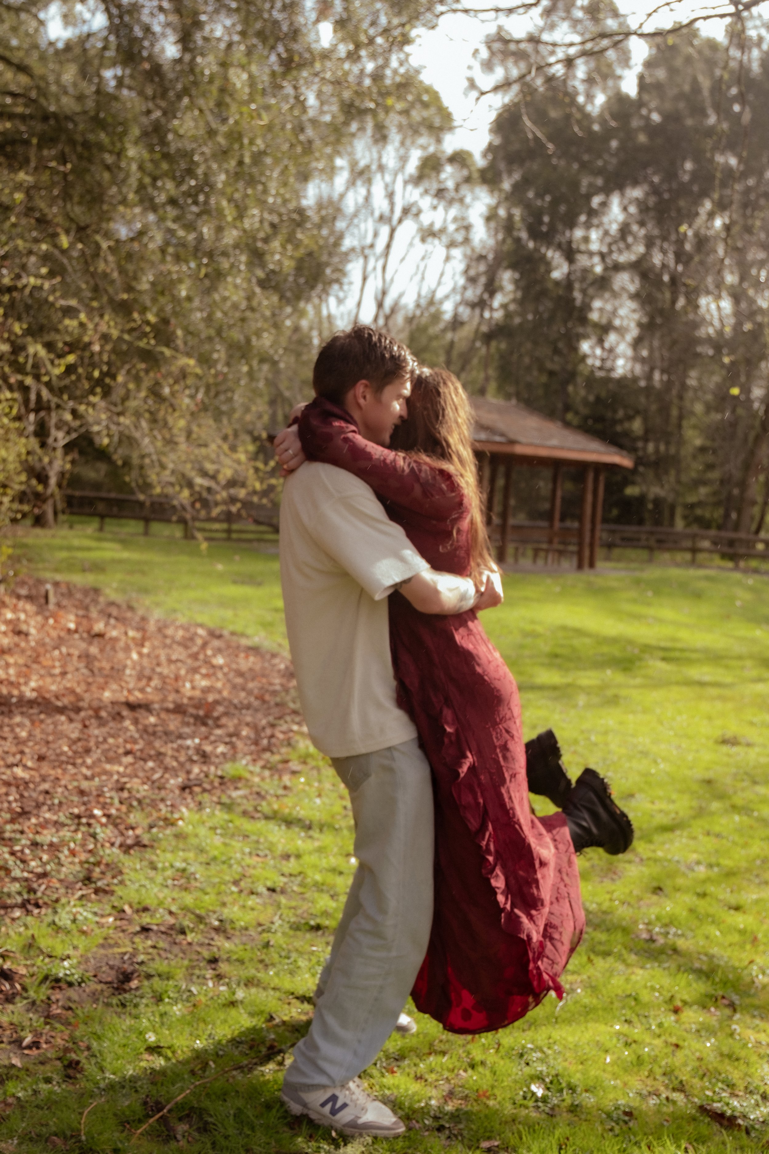 A young man lifting a young woman in a park with trees and a pavilion in the background. Couples photography Hamilton New Zealand. Engagement photography Hamilton New Zealand. Whangarei New Zealand Couples photographer