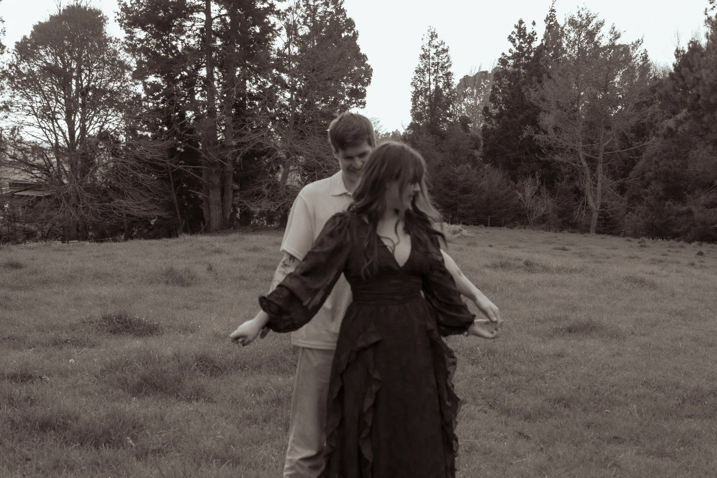 A young man and woman standing outdoors in a grassy field with trees in the background. The woman is wearing a long dress with puffy sleeves, and the man is dressed casually in a light-colored shirt and pants. Engagement photography Hamiton. 