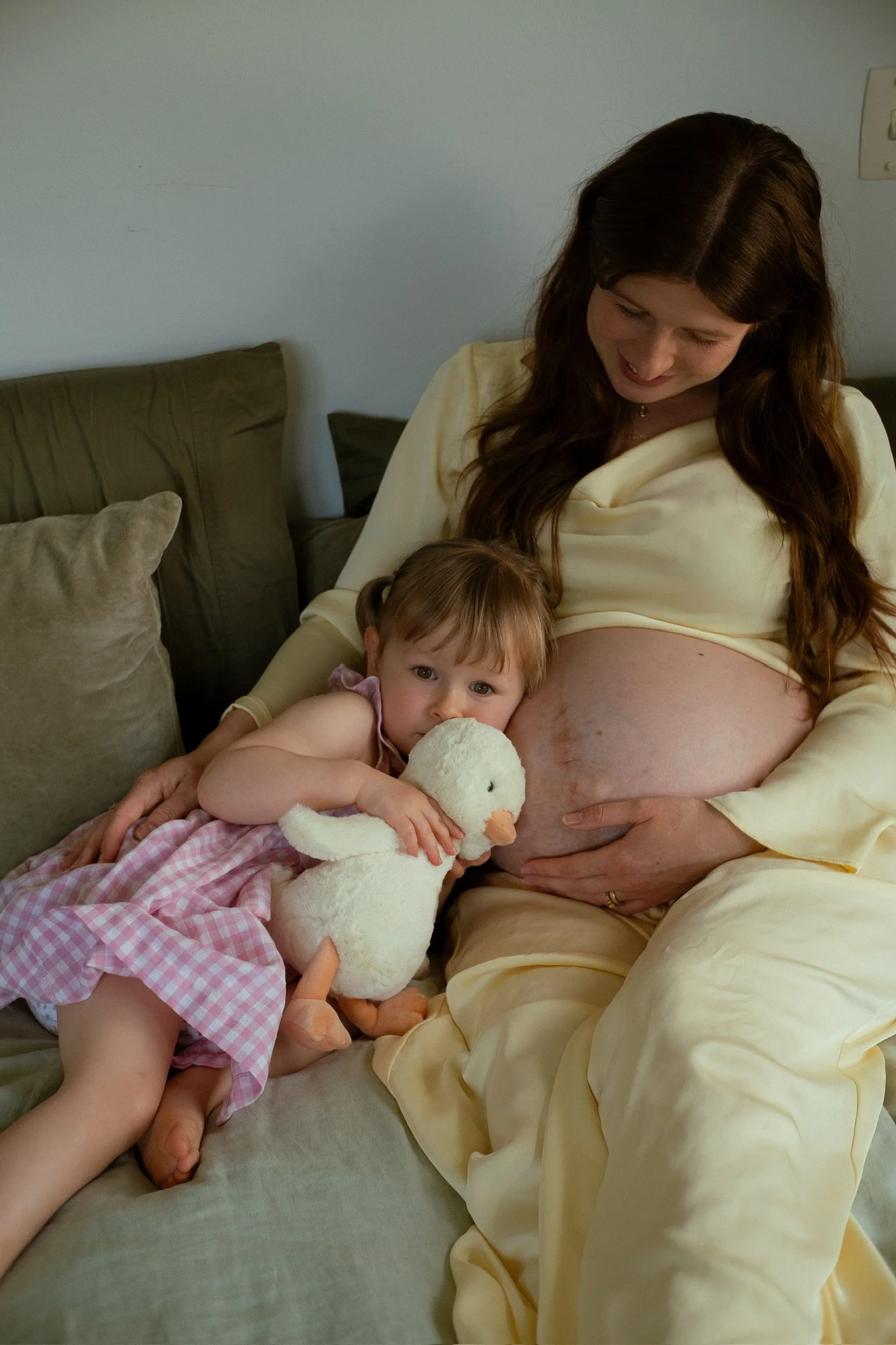 A pregnant woman with long dark hair sitting on a beige couch, smiling and looking down at her belly. A young girl in a pink dress is sitting close to her, hugging a stuffed duck toy and resting her head on the woman's belly, looking at the camera.