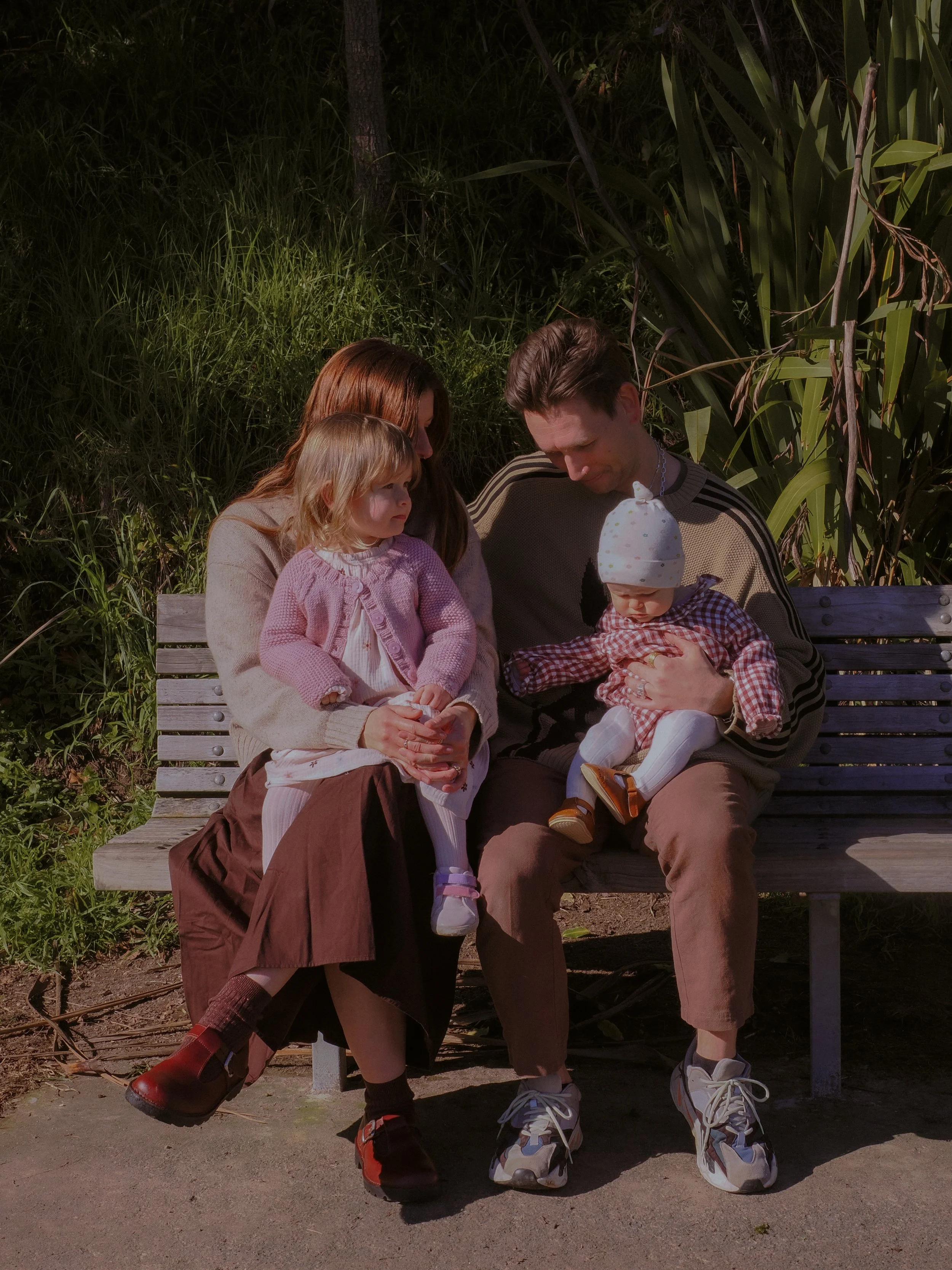 A family of four sitting on a park bench, with a woman, two young children, and a man, surrounded by green foliage. Family Photography Waikato New Zealand. Family Photography Hamilton New Zealand. Northland Wedding Photography