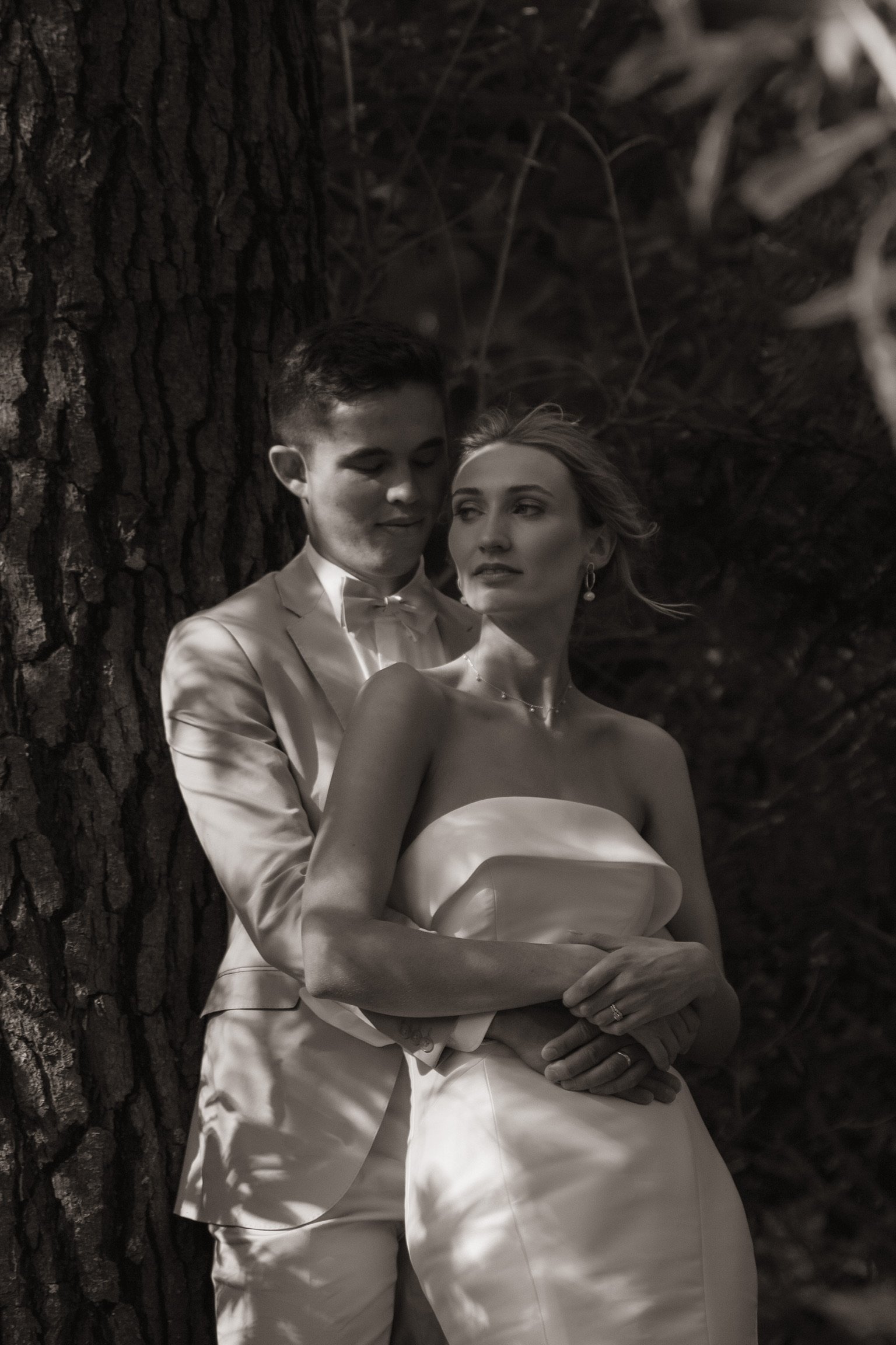 A black-and-white photo of a couple in wedding attire, standing outdoors by a tree, with the groom embracing the bride from behind.  Wedding Photography Waikato New Zealand. Wedding Photography Hamilton New Zealand. Northland Wedding Photography