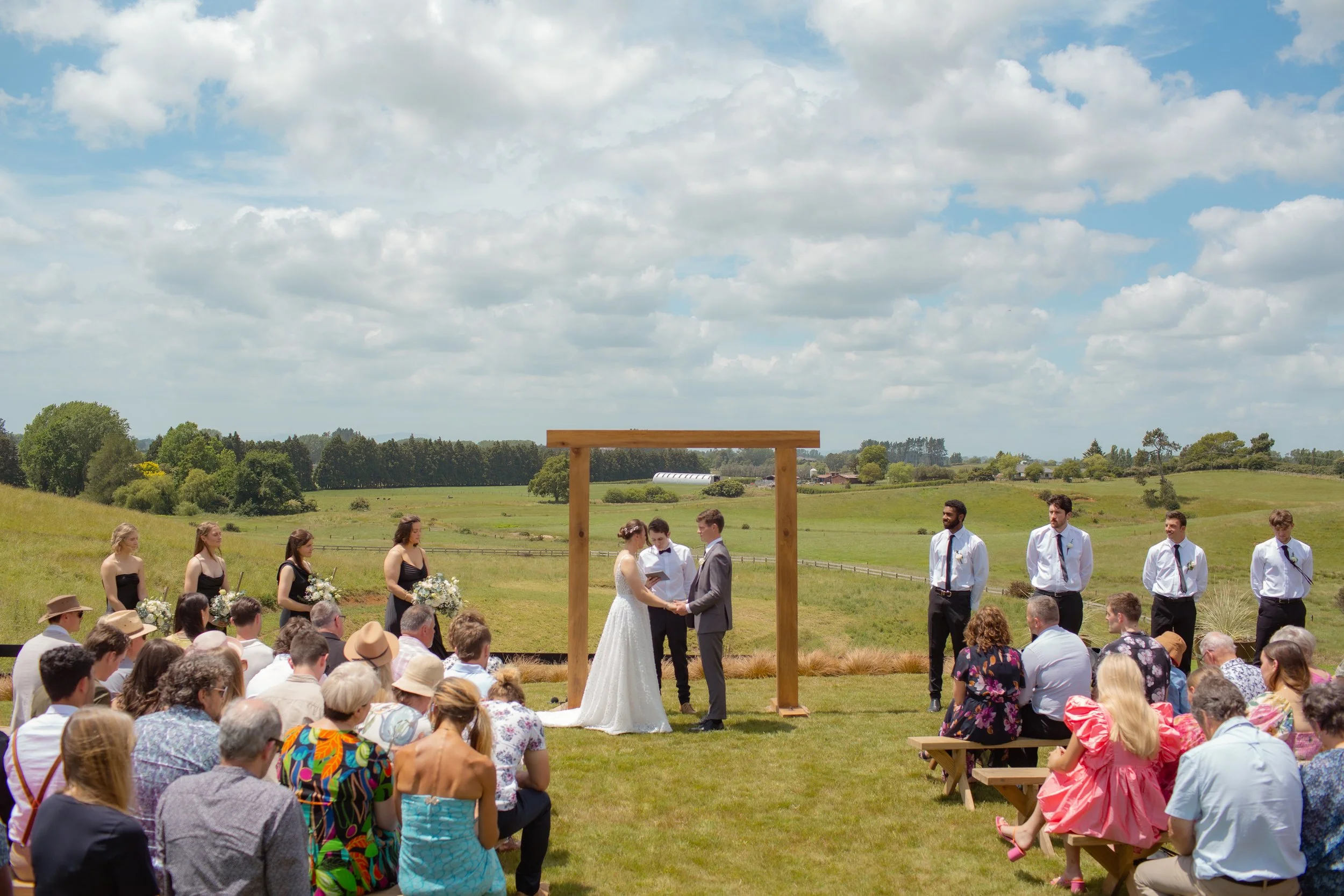 Outdoor wedding ceremony with bride and groom holding hands under a wooden arch. Bridesmaids and groomsmen stand on either side. Guests are seated on benches and chairs, with some wearing colorful or patterned clothing. The background features green 