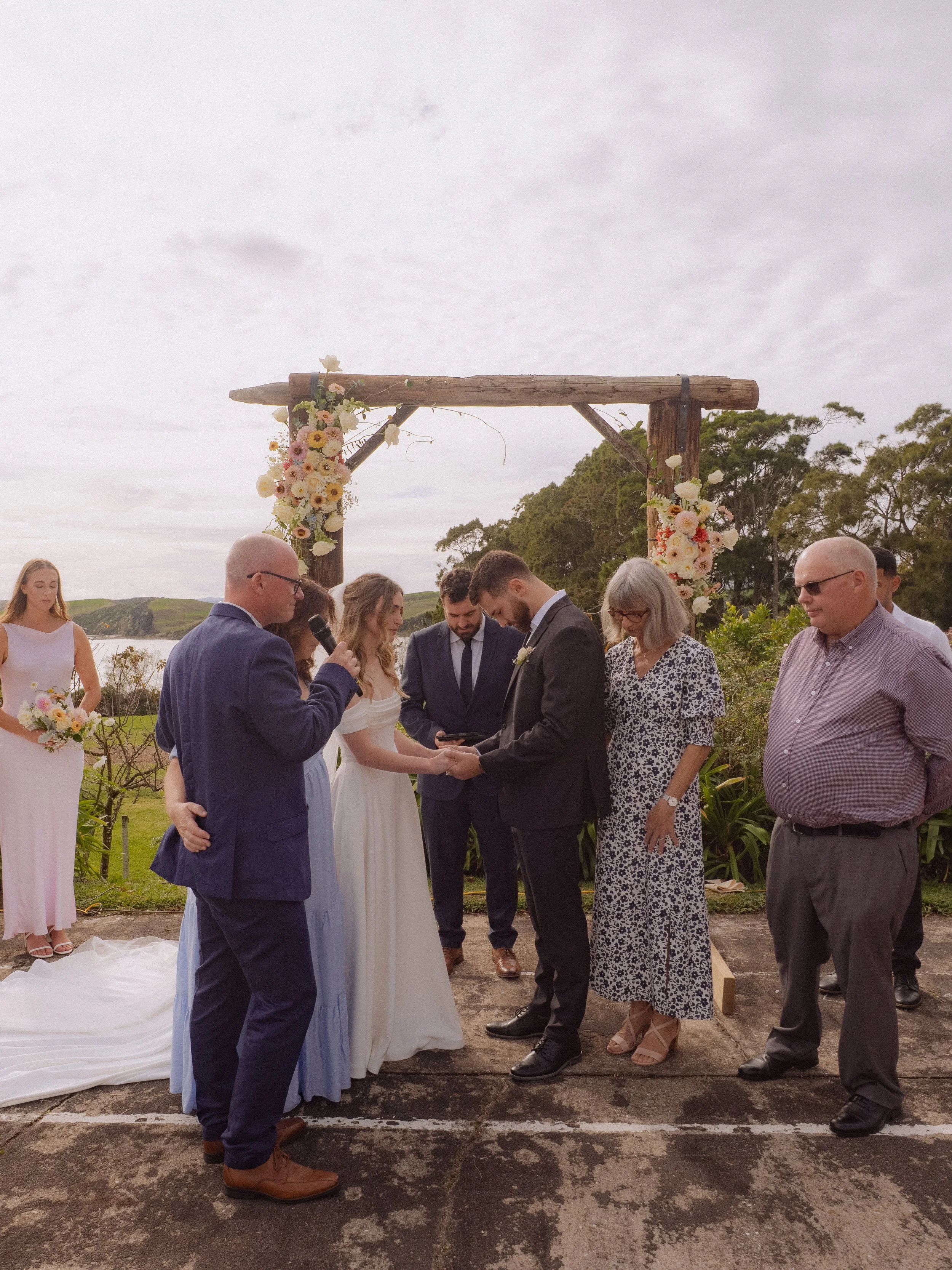 Wedding ceremony outdoors with couple holding hands under a wooden arch decorated with flowers, surrounded by friends and family.  Wedding Photography Waikato New Zealand. Wedding Photography Hamilton New Zealand. Northland Wedding Photography
