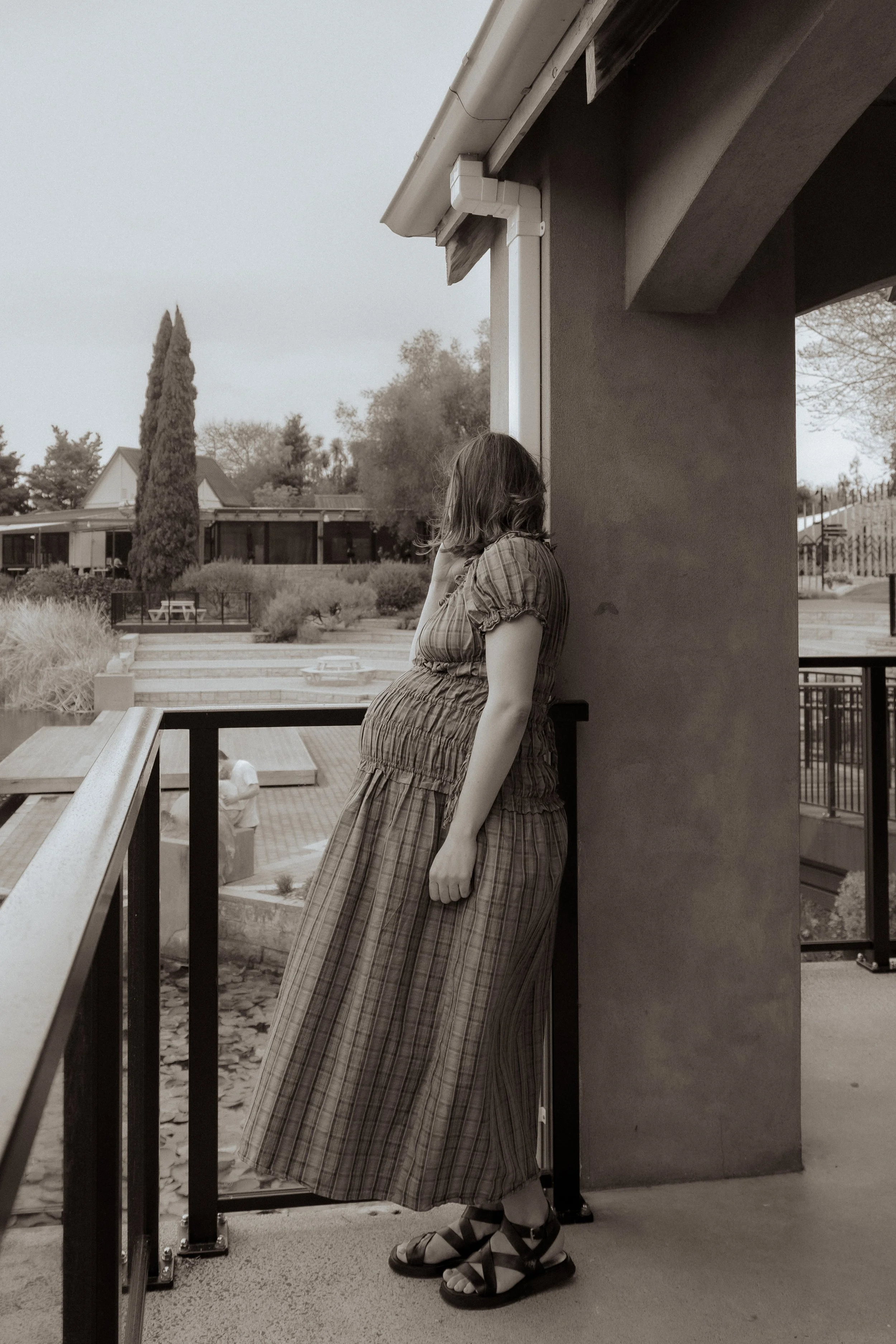 A woman in a plaid dress standing on a balcony, looking away, with her hand touching her face, in black and white. Maternity Photography Hamilton New Zealand