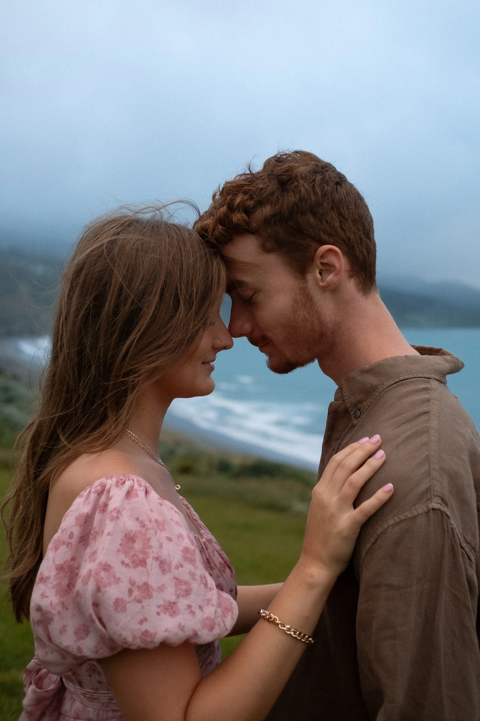 A young couple stands close together with their foreheads touching, outdoors near a body of water, sharing an intimate moment at dusk. Couples photography Raglan New Zealand. Engagement photography Hamilton New Zealand. Whangarei New Zealand Couples 
