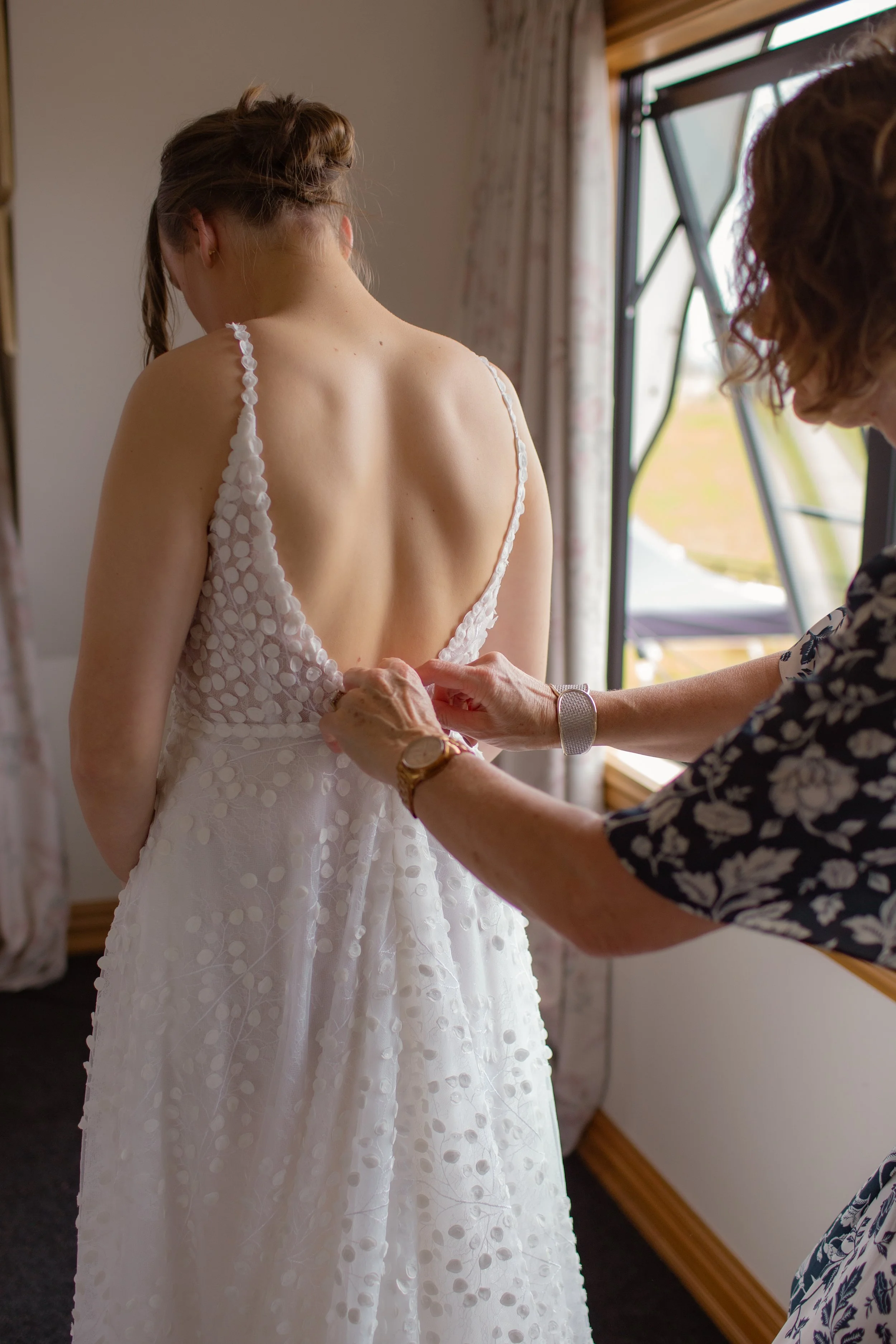A woman is helping a bride put on her wedding dress, with the bride standing by a window.  Wedding Photography Waikato New Zealand. Wedding Photography Hamilton New Zealand. Northland Wedding Photography