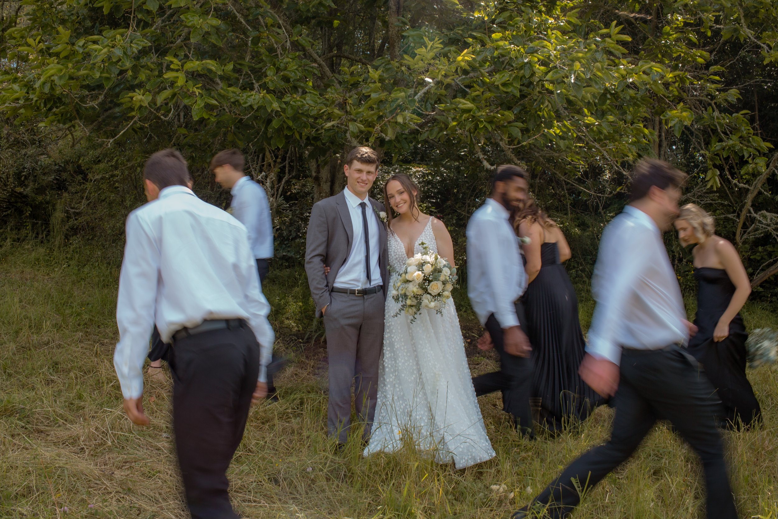 A bride and groom standing in the center, smiling, surrounded by wedding guests walking in a natural outdoor setting with green trees and grass.  Wedding Photography Waikato New Zealand. Wedding Photography Hamilton New Zealand. Northland Wedding 