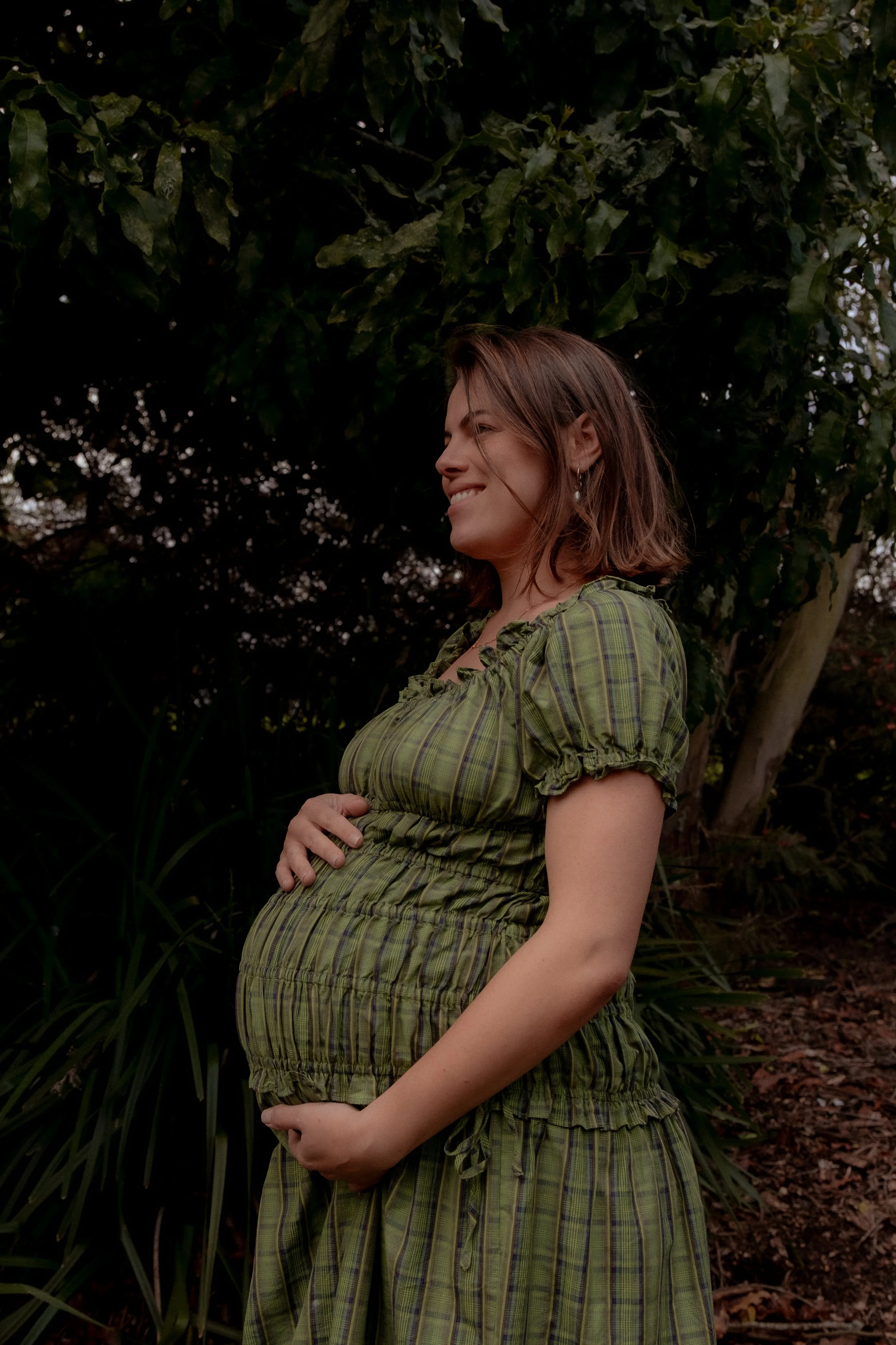 A pregnant woman smiling outdoors, holding her belly with her right hand, in front of green foliage. Maternity photography Hamilton New Zealand. Family photographer