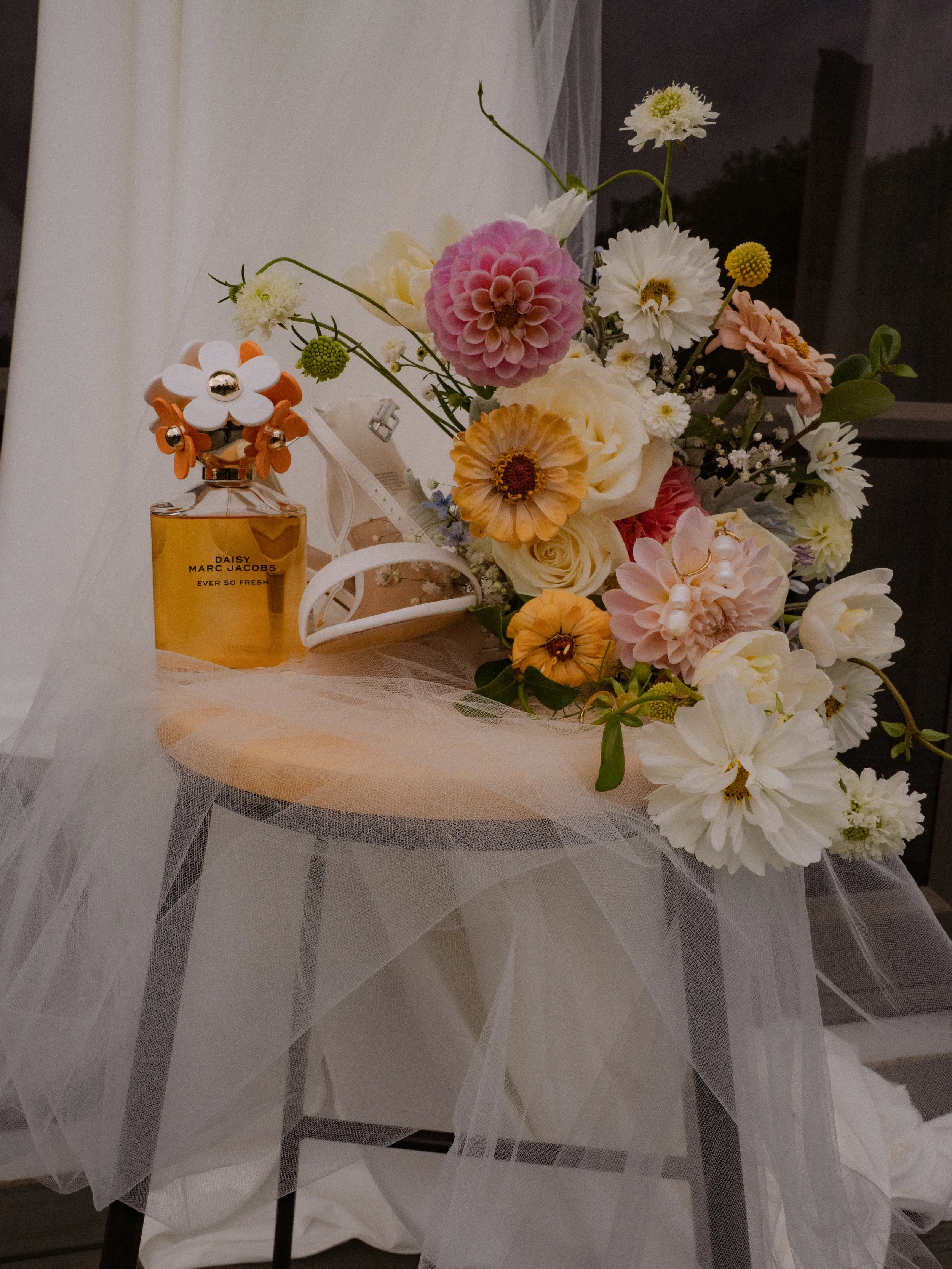 A table decorated with a large bouquet of mixed flowers, a perfume bottle labeled Daisy by Marc Jacobs, and a white high-heeled shoe, all covered partly by a sheer fabric.  Wedding Photography Waikato New Zealand. Wedding Photography Hamilton 