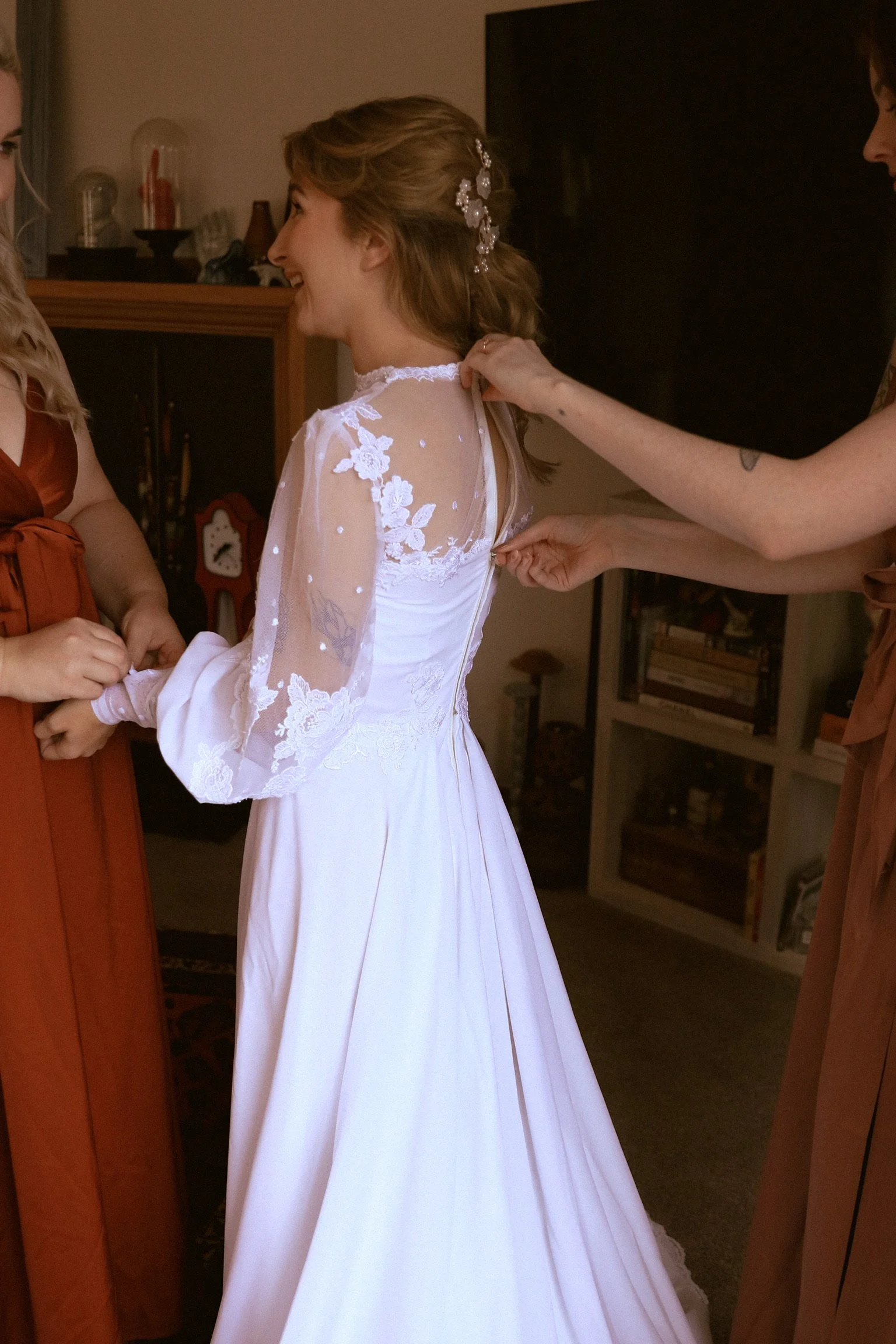 A bride wearing a white wedding dress with lace and floral details is staying as someone helps her adjust her dress, surrounded by two women in brown dresses. Wedding Photography Waikato New Zealand. Wedding Photography Hamilton New Zealand
