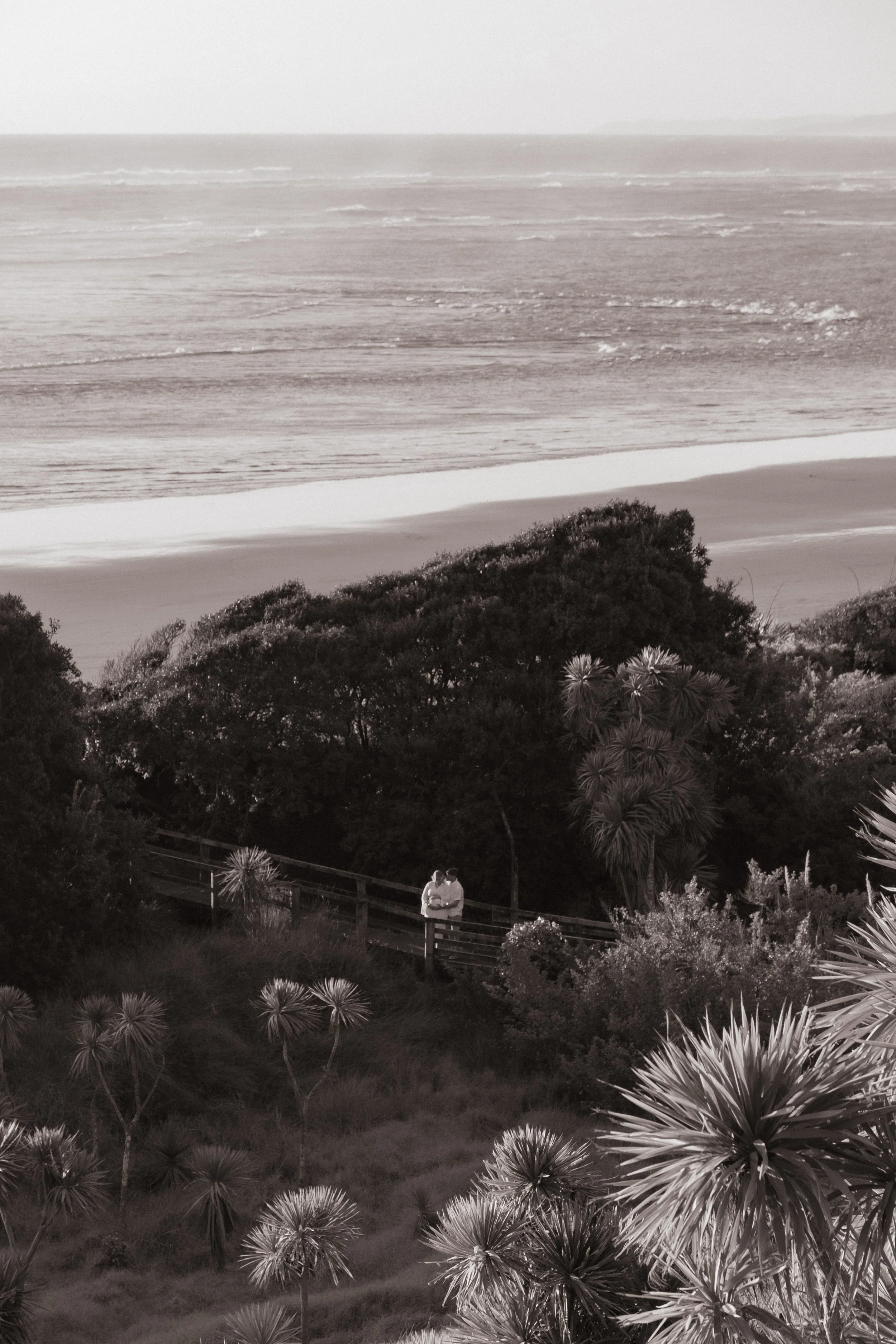 Couple standing on a boardwalk overlooking a beach with waves and sand, surrounded by trees and plants. Raglan New Zealand