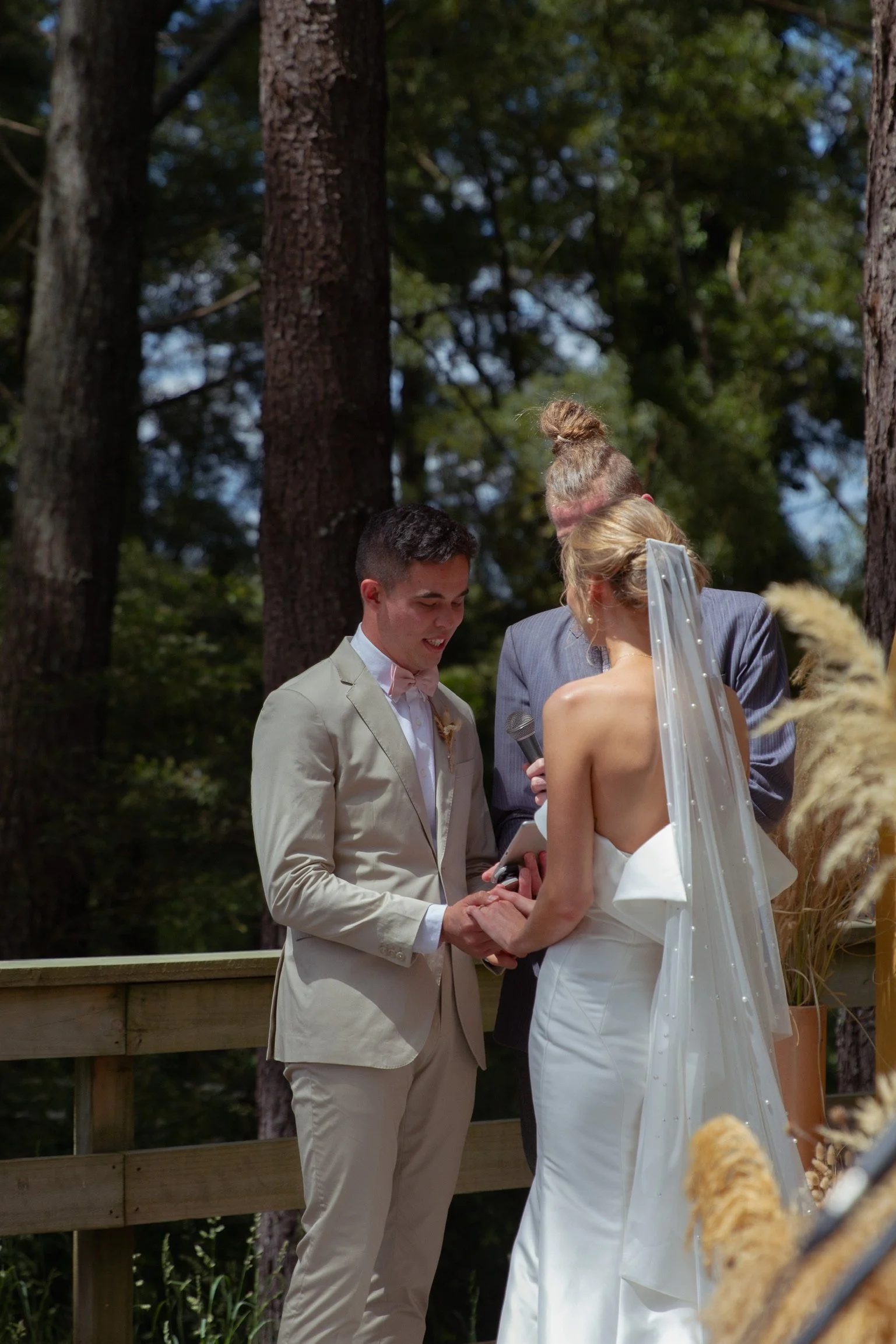A wedding ceremony taking place outdoors in a wooded area with a bride and groom exchanging vows, officiant holding a mic, and lush trees in the background.  Wedding Photography Waikato New Zealand. Wedding Photography Hamilton New Zealand. 
