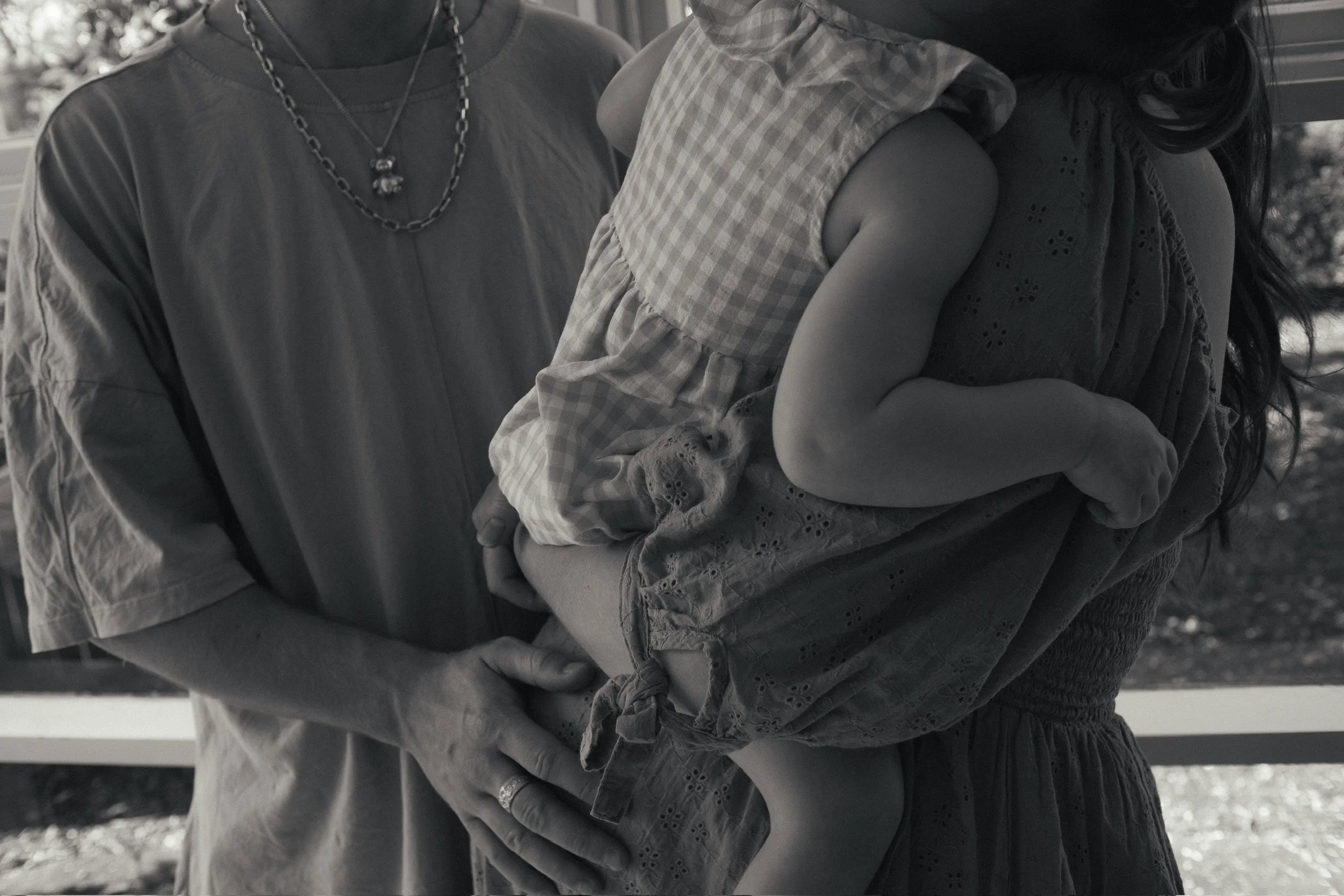 A woman is holding a young girl in her arms. The woman wears a necklace and a loose shirt, while the child is in a checkered dress. The scene is in black and white. Maternity Photography. Family Photographer. Hamilton New Zealand Photography