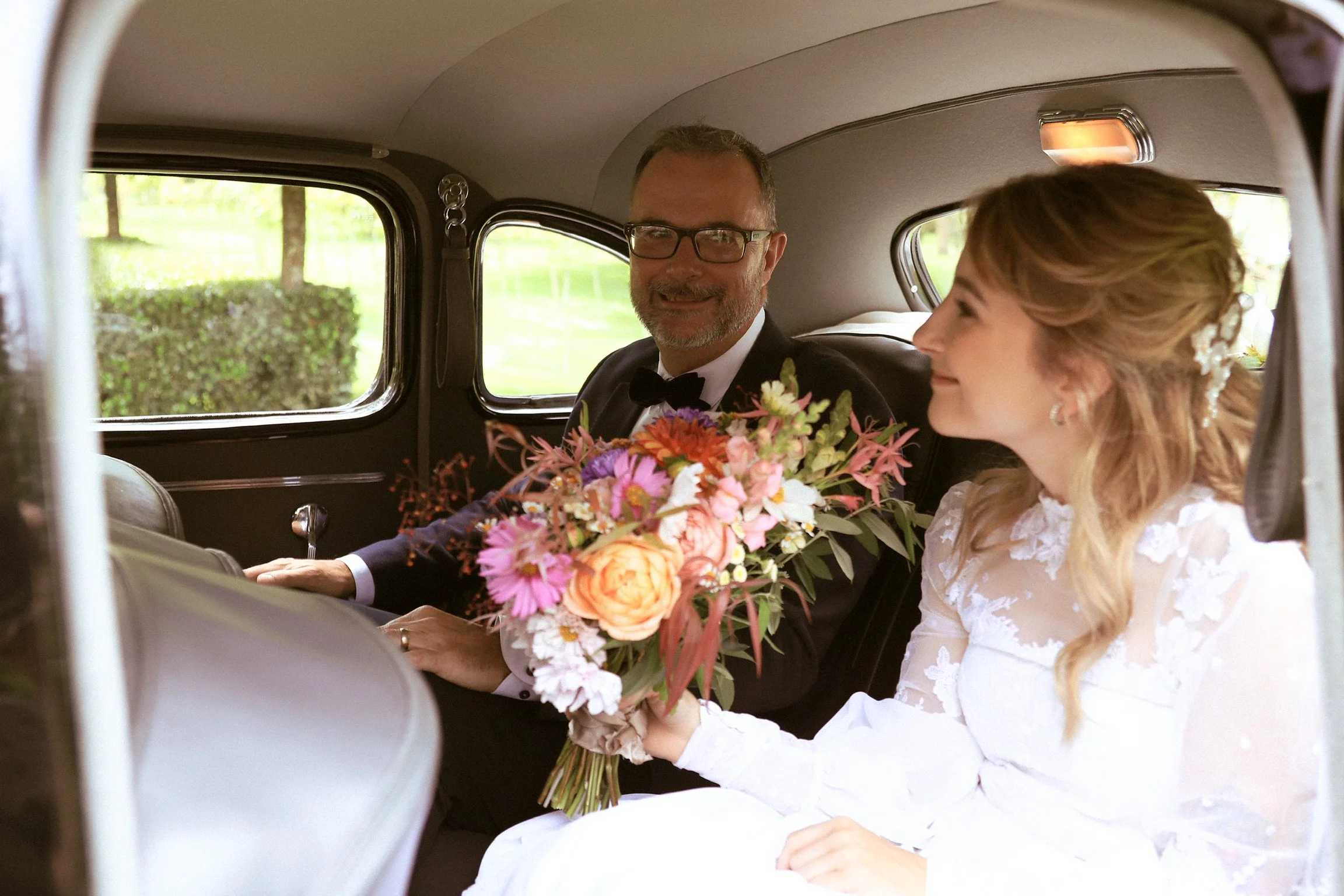 A bride and groom sitting inside a vintage car, with the bride holding a large bouquet of colorful flowers, smiling and looking at each other. Wedding Photography Waikato New Zealand. Wedding Photography Hamilton New Zealand