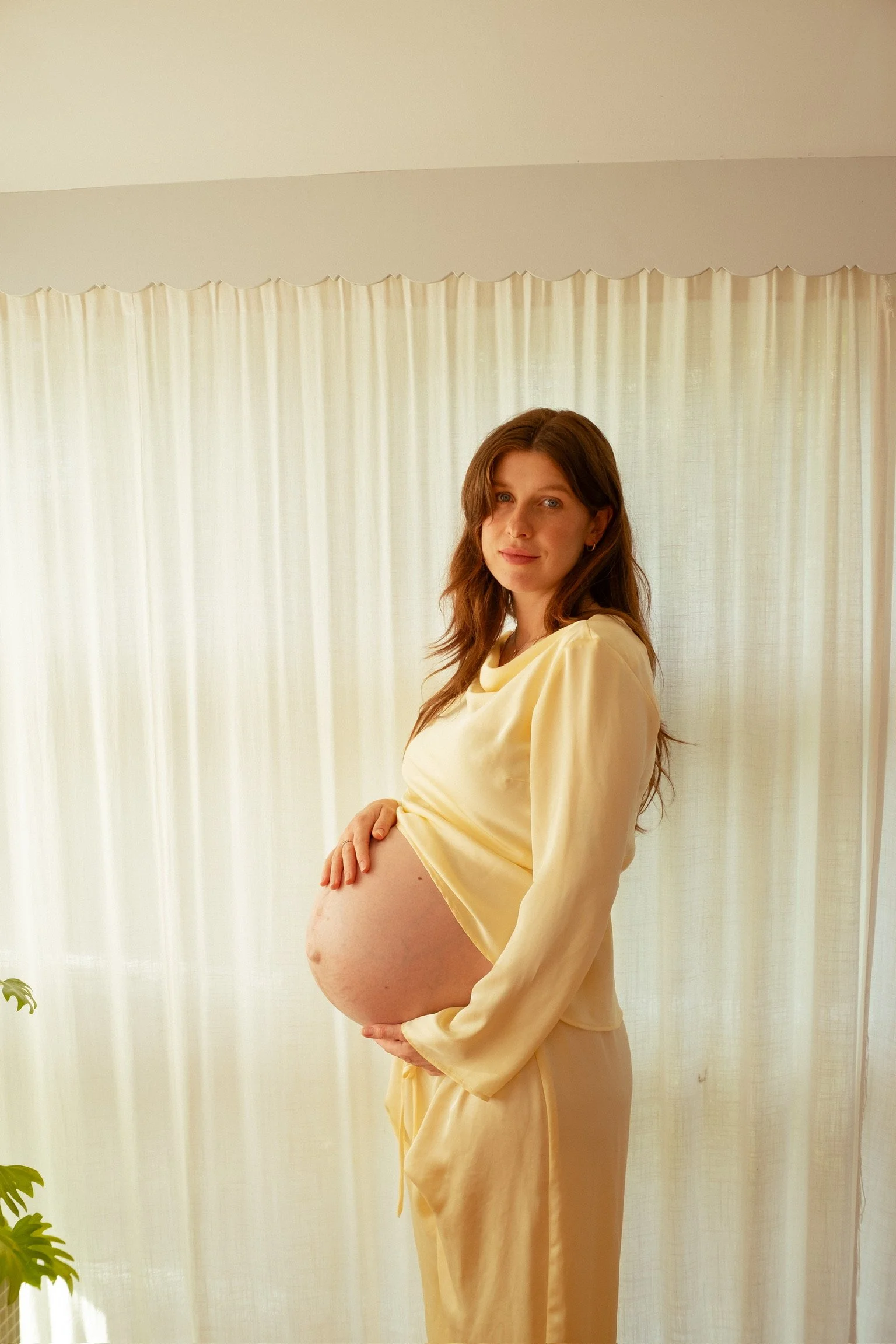 Pregnant woman in cream-colored pajamas standing indoors in front of light-colored curtains, holding her belly and smiling gently. Maternity photography. Hamilton New Zealand Photographer