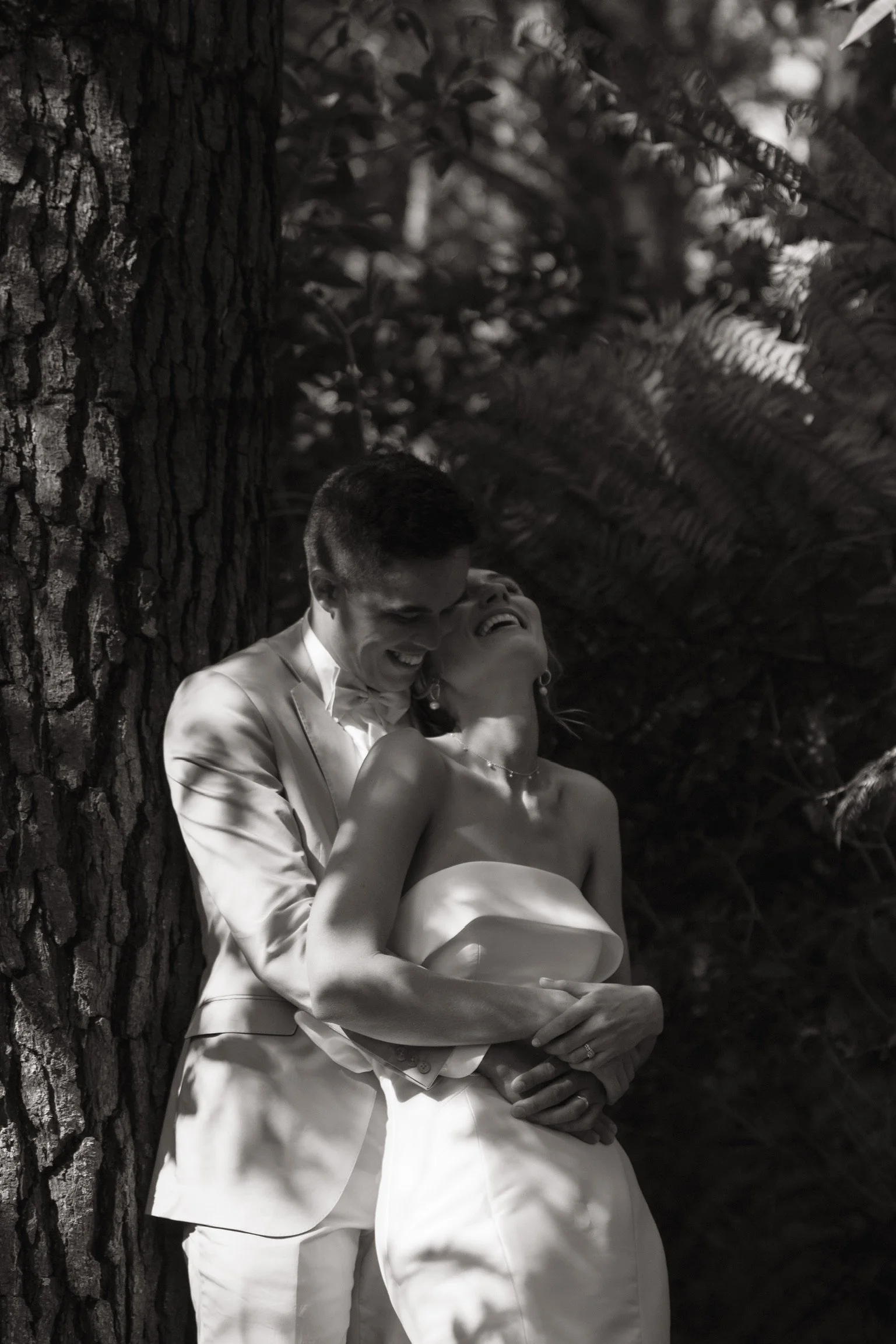 Black-and-white photo of a happy couple in wedding attire embracing and laughing in front of a large tree trunk and foliage.