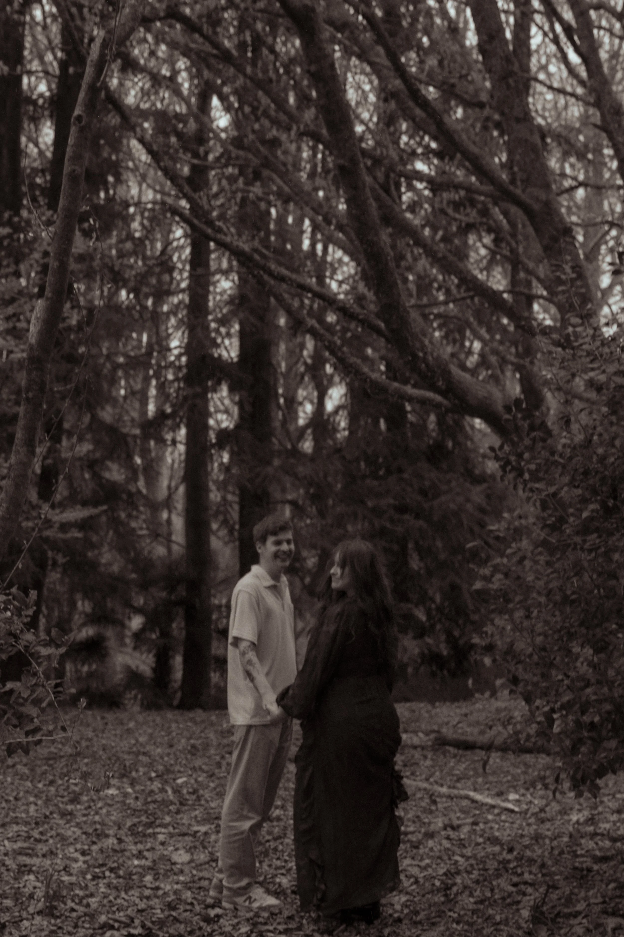 A young man and woman holding hands and smiling in a wooded forest with tall trees and leaf-covered ground. Couples photography Hamilton New Zealand. Engagement photography Hamilton New Zealand. Whangarei New Zealand Couples photographer