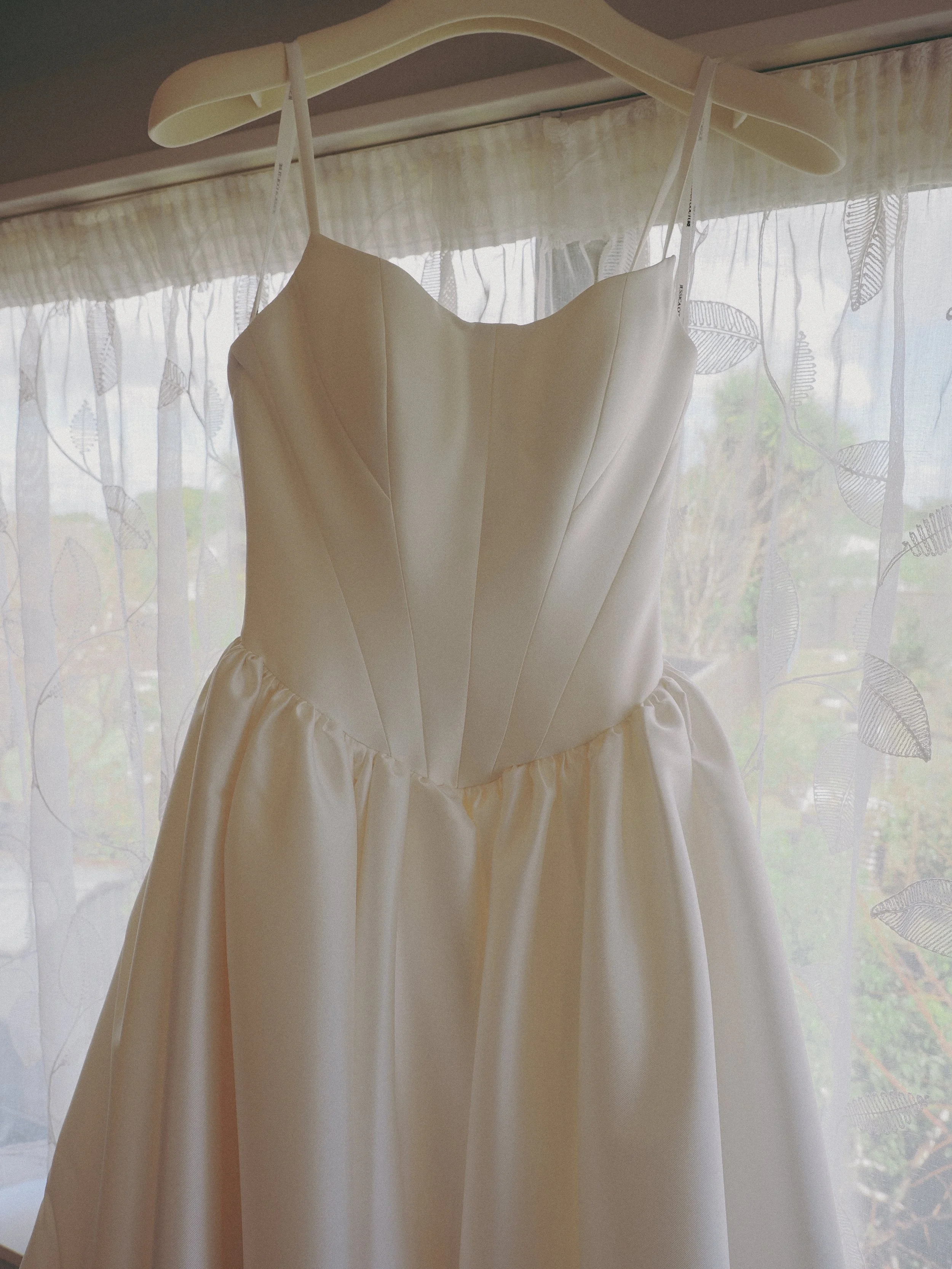 A wedding dress hanging on a hanger in front of a window with sheer curtains featuring a leaf pattern.