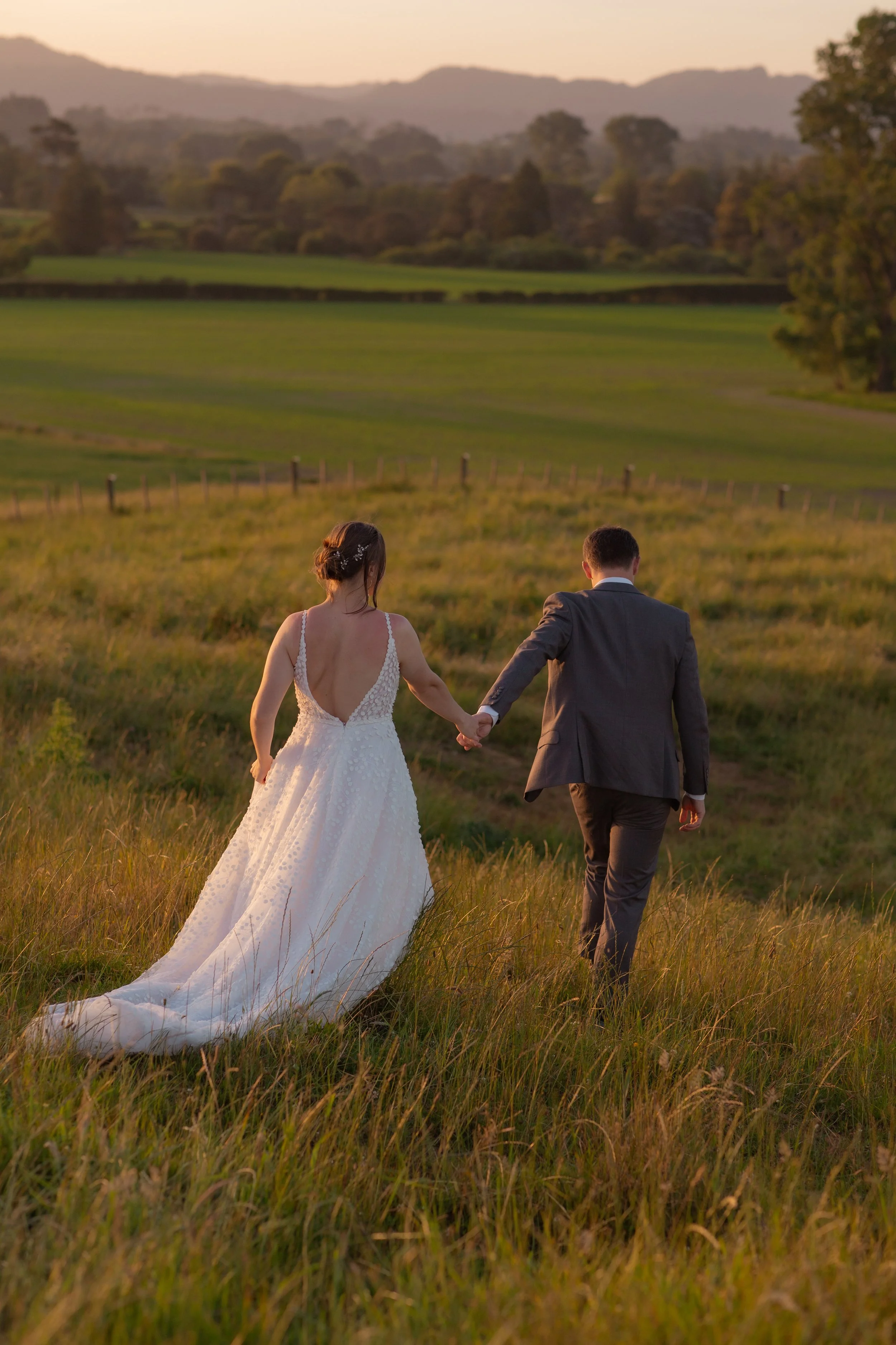A bride in a white wedding dress and a groom in a gray suit holding hands and walking through a grassy field at sunset with rolling hills and trees in the background.