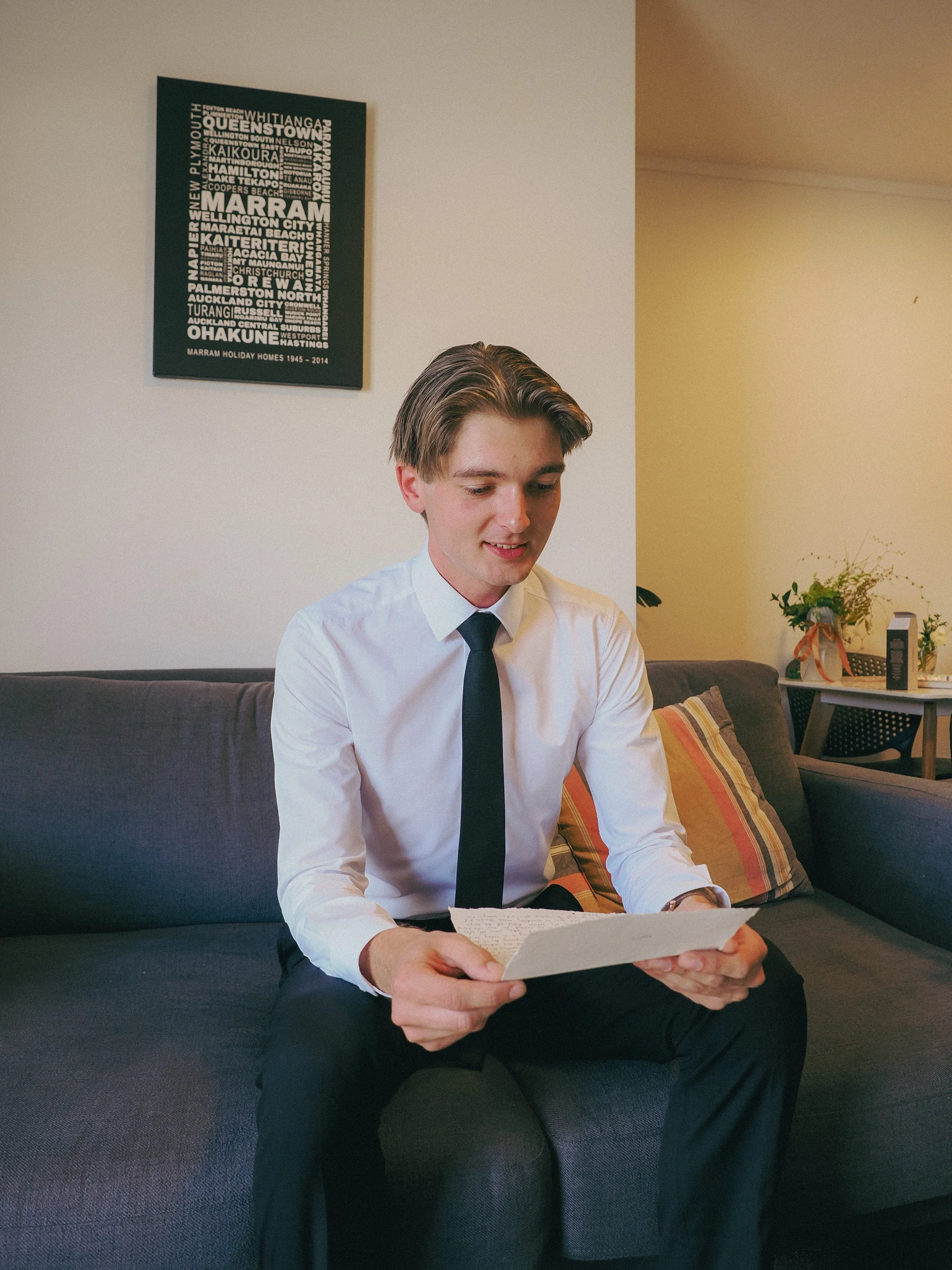 A young man dressed in a white shirt and dark tie sitting on a dark gray couch, reading a handwritten letter or note, with a beige and orange striped pillow behind him. A painting of city names and a potted plant are visible in the background.