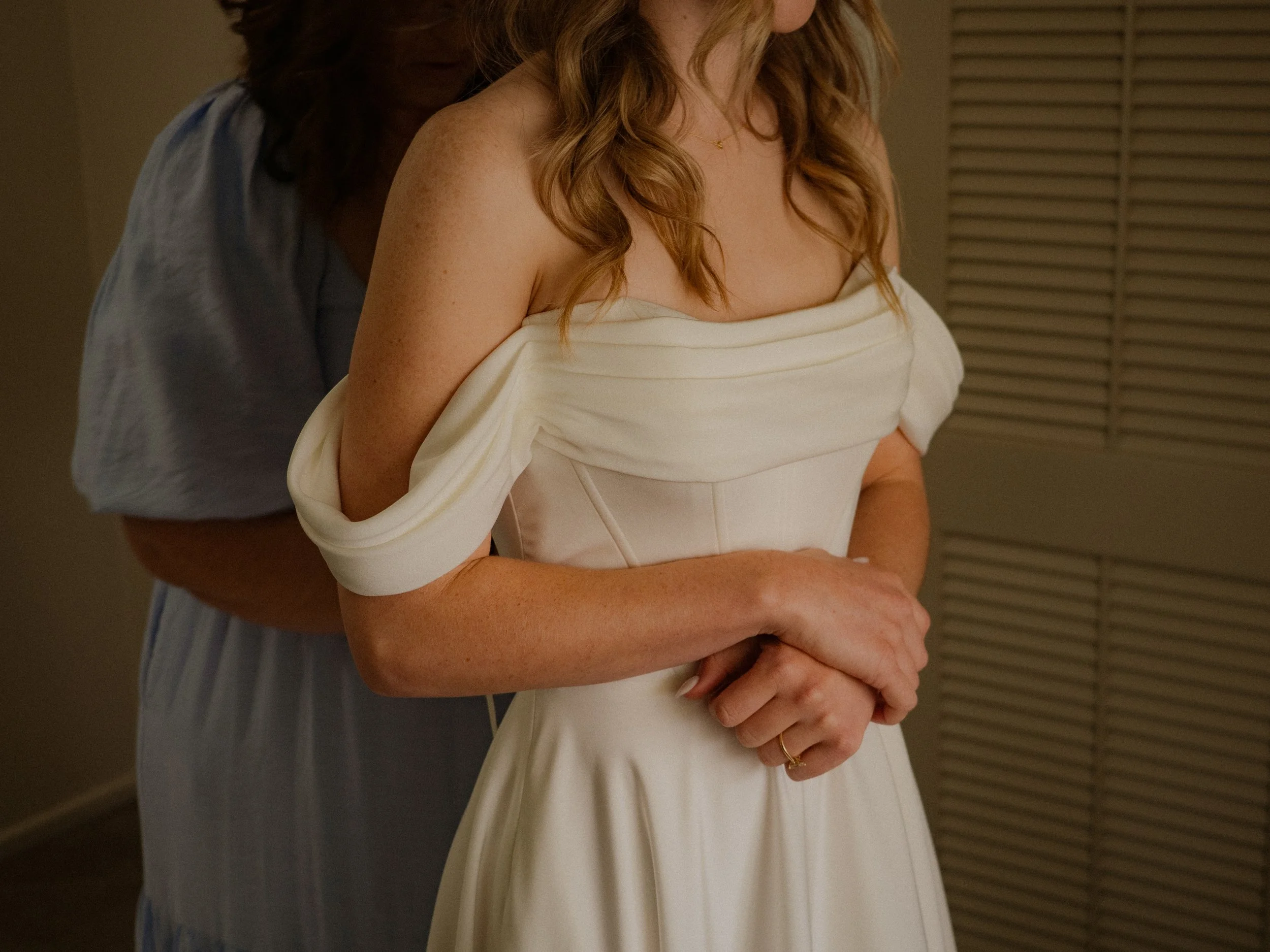 A woman in a white dress with off-the-shoulder sleeves is being assisted from behind by another person, who is helping her dress.  Wedding Photography Waikato New Zealand. Wedding Photography Hamilton New Zealand. Northland Wedding Photography