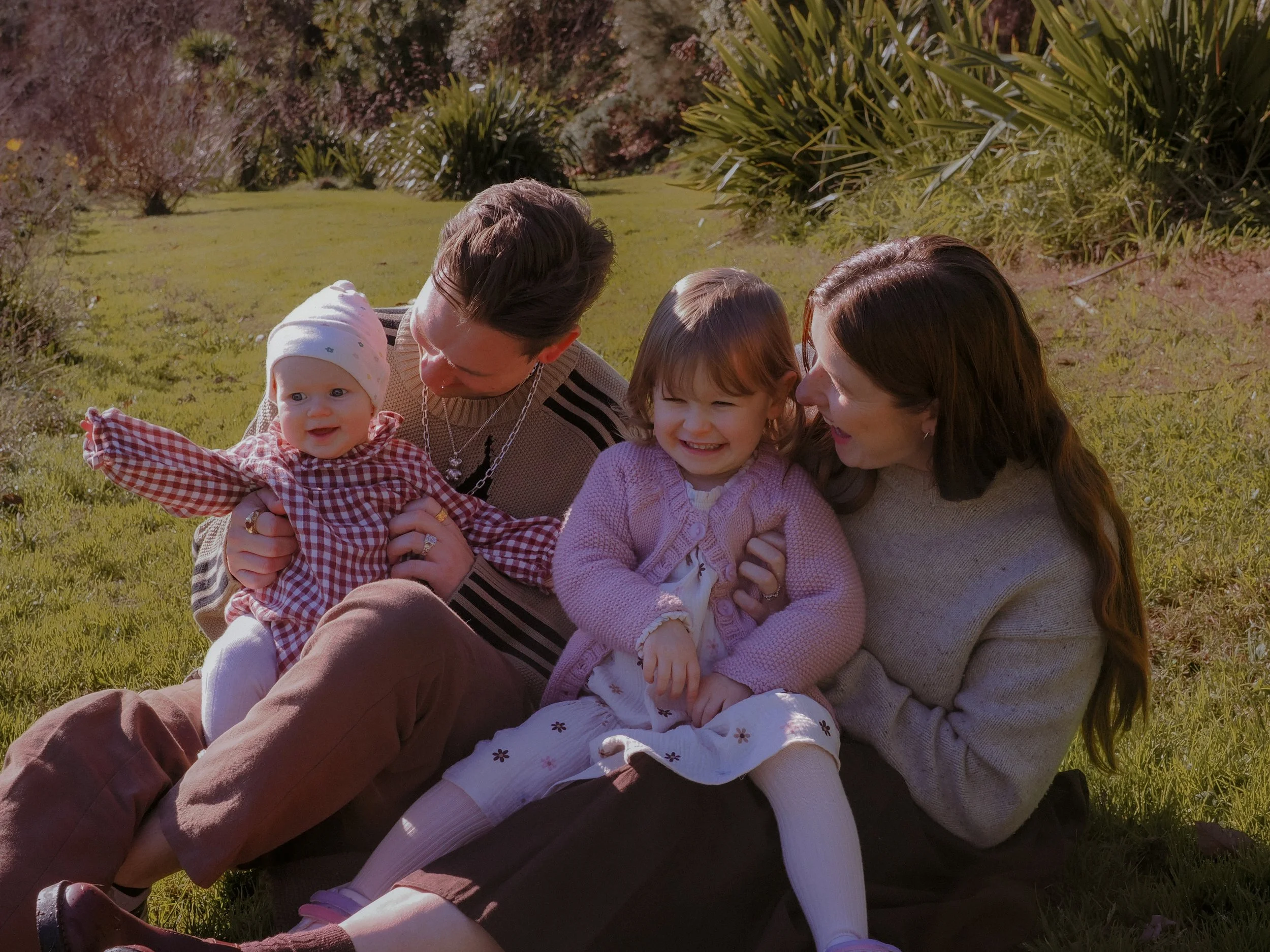 A family of four sitting on grass outdoors, smiling and enjoying time together. Family Photography Waikato New Zealand. Family Photography Hamilton New Zealand. Northland Wedding Photography