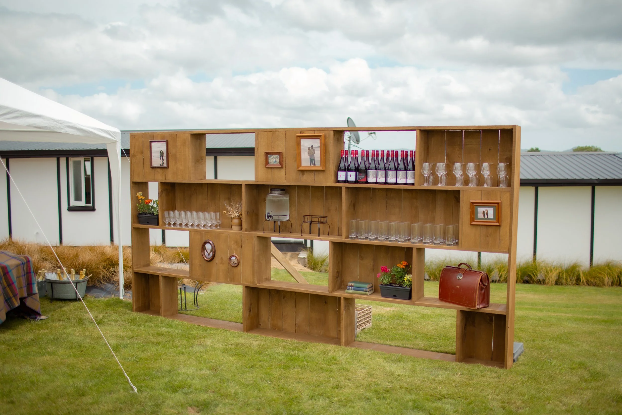 Wooden outdoor bar with glasses, wine bottles, framed photos, a plant, books, and a brown leather bag, set up on a lawn with a tent and buildings in the background.  Wedding Photography Waikato New Zealand. Wedding Photography Hamilton New Zealand. 