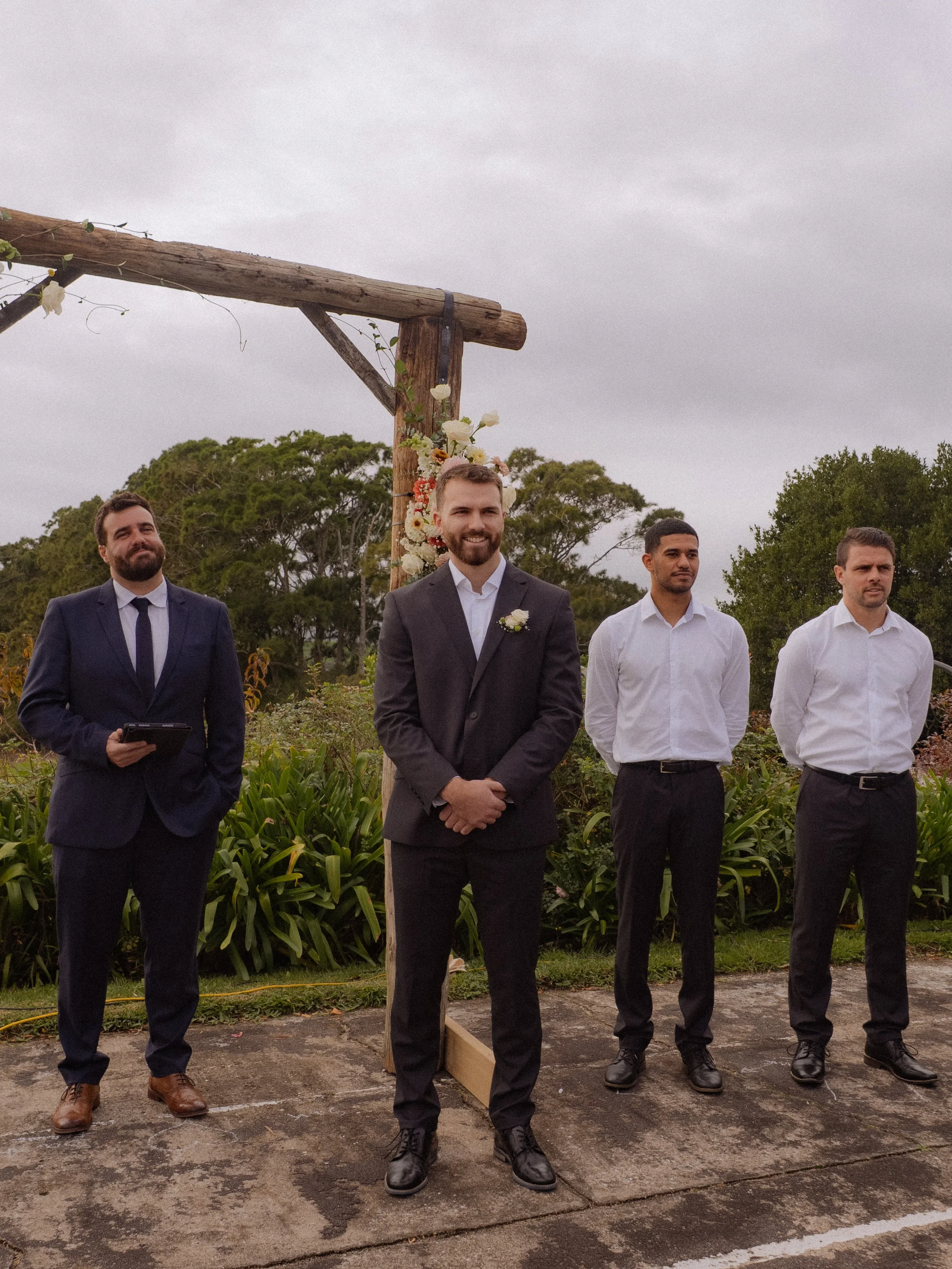 Groom and three groomsmen standing outdoors near a wooden wedding arch decorated with flowers, under cloudy sky.  Wedding Photography Waikato New Zealand. Wedding Photography Hamilton New Zealand. Northland Wedding Photography