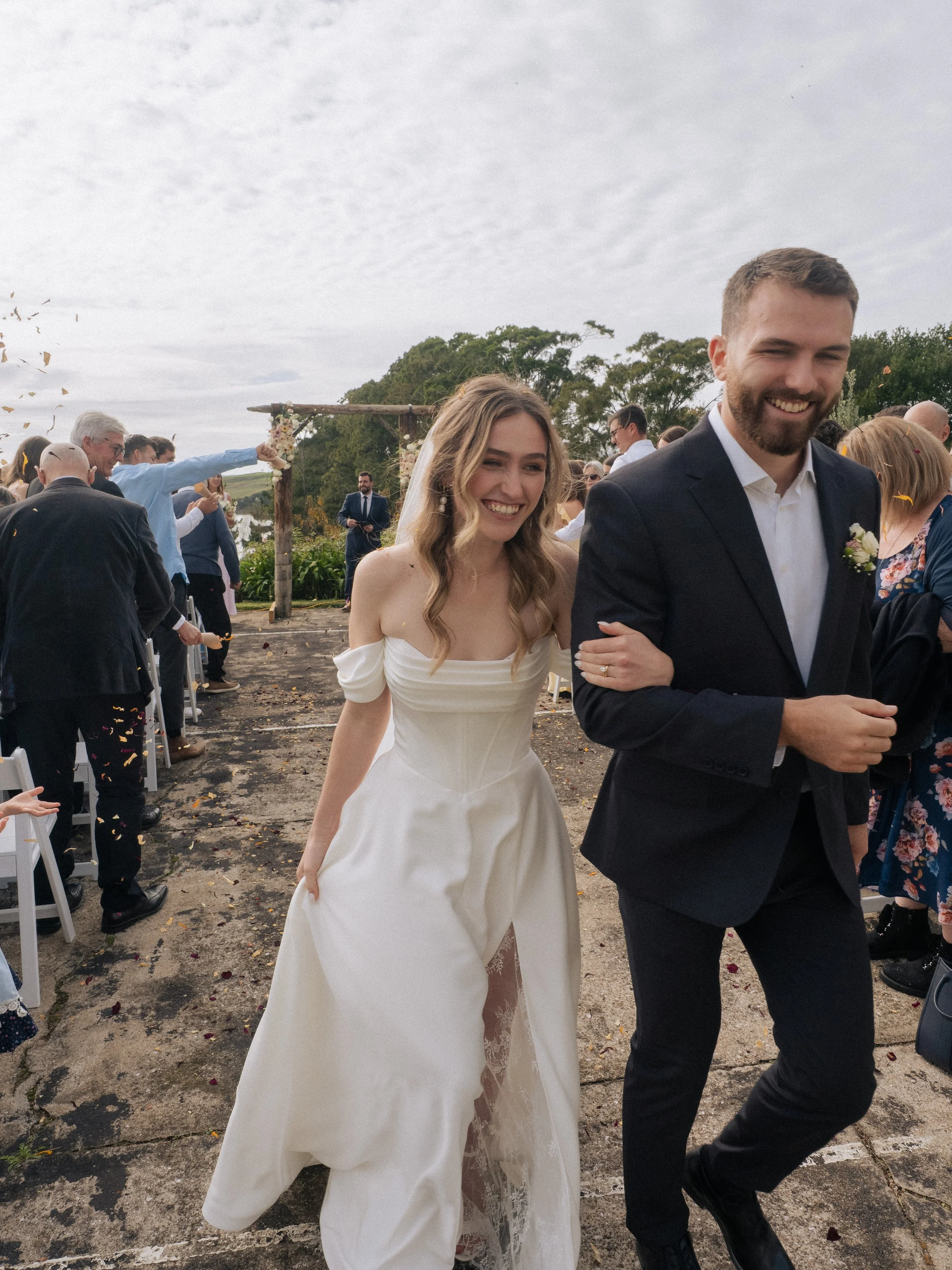 Bride in a white wedding dress walking arm in arm with groom in a dark suit at an outdoor wedding ceremony, guests in background celebrating.  Wedding Photography Waikato New Zealand. Wedding Photography Hamilton New Zealand. Northland Wedding Photo