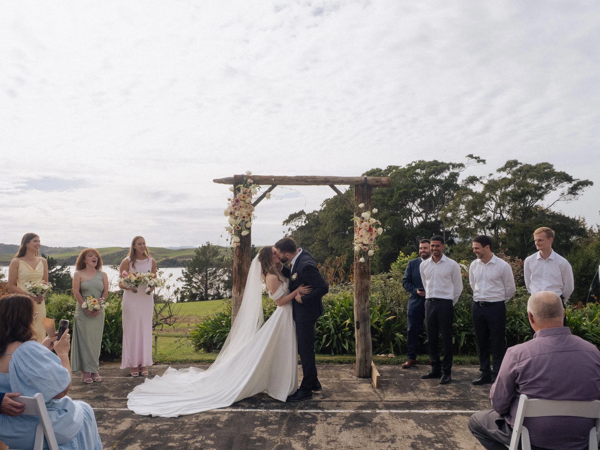A wedding ceremony outdoors with a bride and groom kissing under a wooden arch decorated with flowers, surrounded by bridesmaids and groomsmen, with guests seated nearby and a scenic lake and trees in the background.  Wedding Photography Waikato 