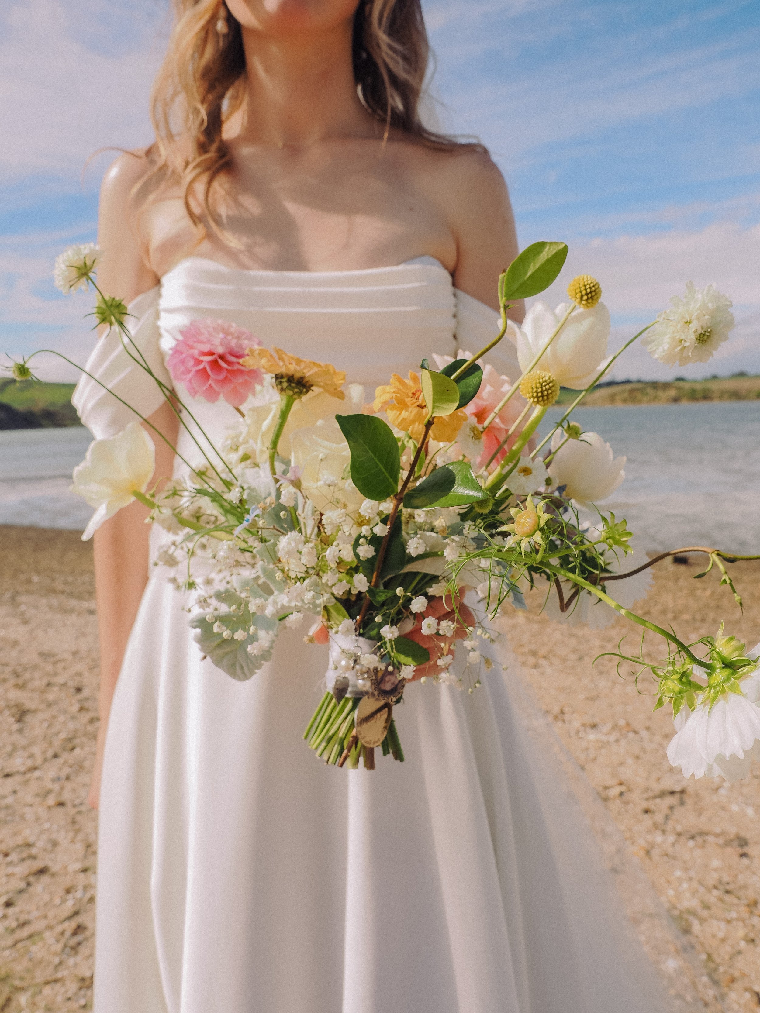 A woman in a white dress holding a bouquet of mixed flowers on a beach with water and blue sky in the background.  Wedding Photography Waikato New Zealand. Wedding Photography Hamilton New Zealand. Northland Wedding Photography