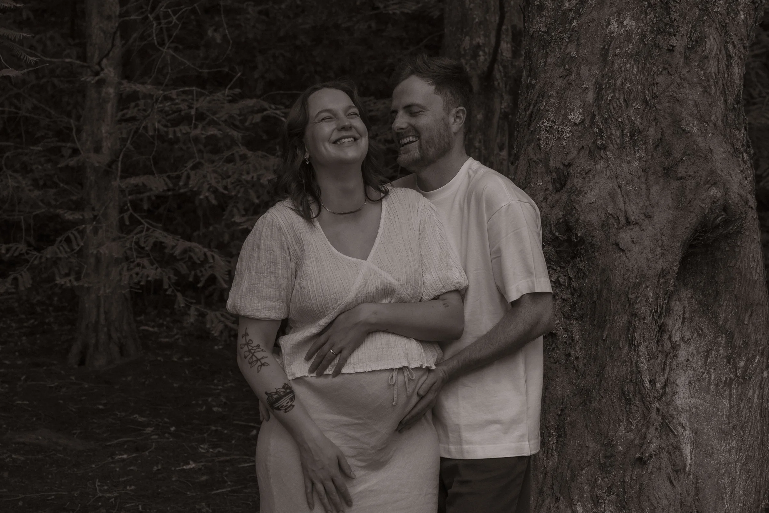 A man and woman smiling and holding each other, standing outdoors near a large tree in a forested area. Maternity Photography. Hamilton New Zealand Photographer