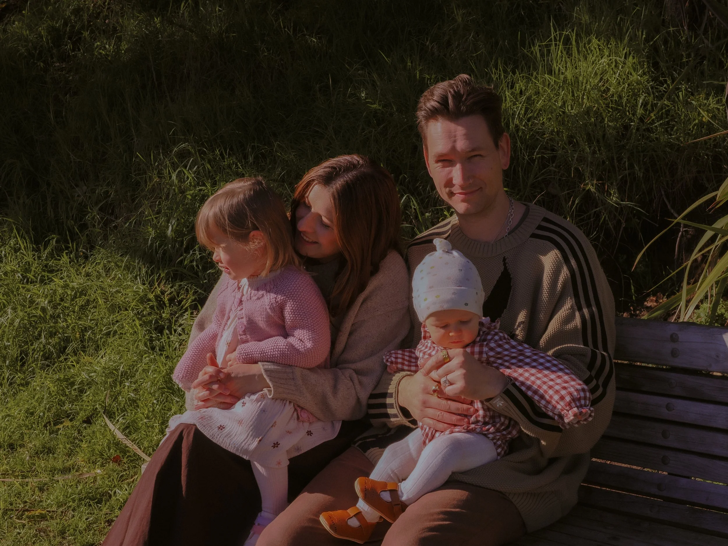 A family sitting on a park bench outdoors, with a young girl, a woman, a man, and a baby girl. The family is enjoying a sunny day surrounded by greenery. Family Photography Waikato New Zealand. Family Photography Hamilton New Zealand. 