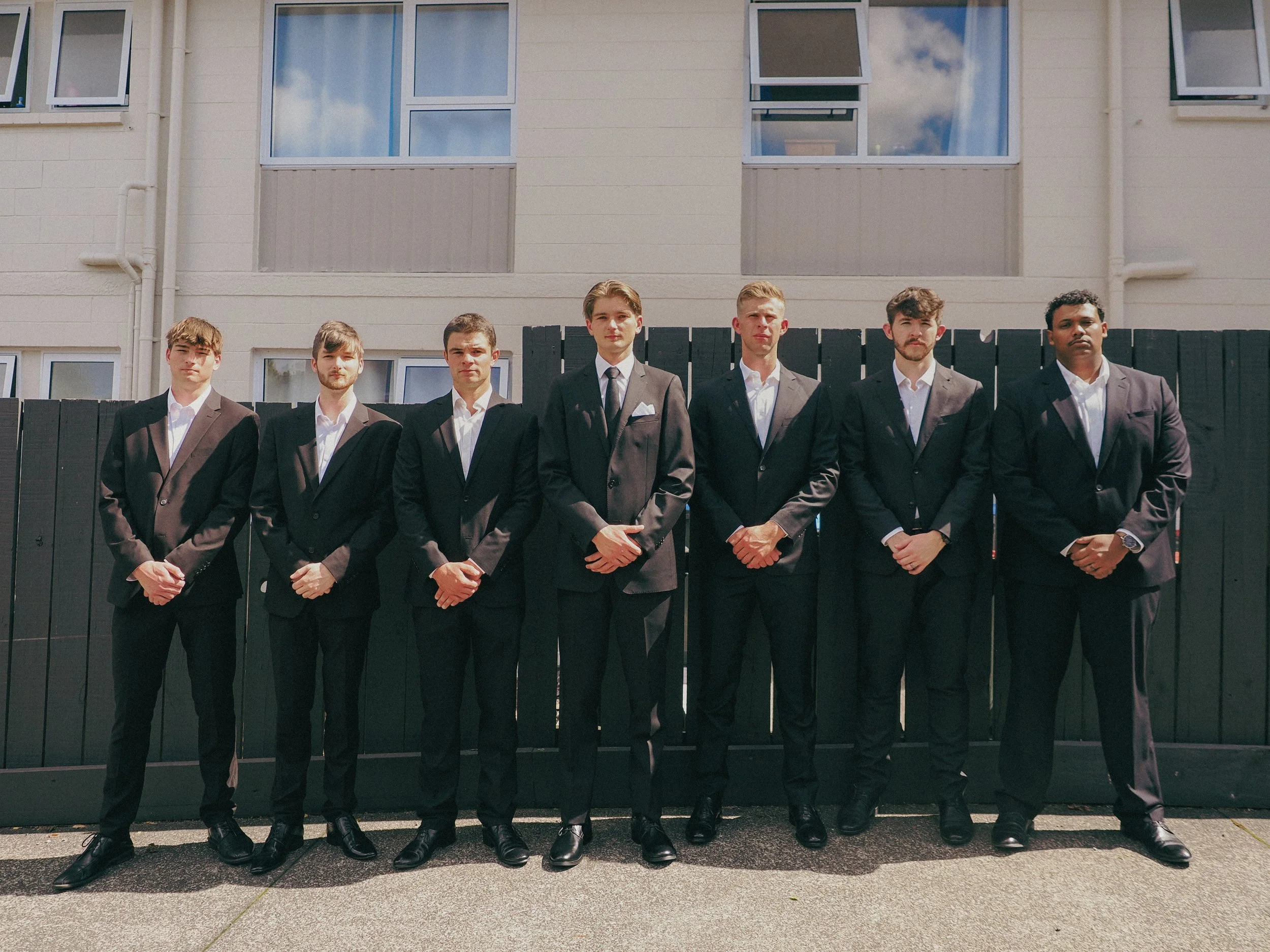 Seven men dressed in formal suits standing in front of a black fence and a modern building, posing for a photo.