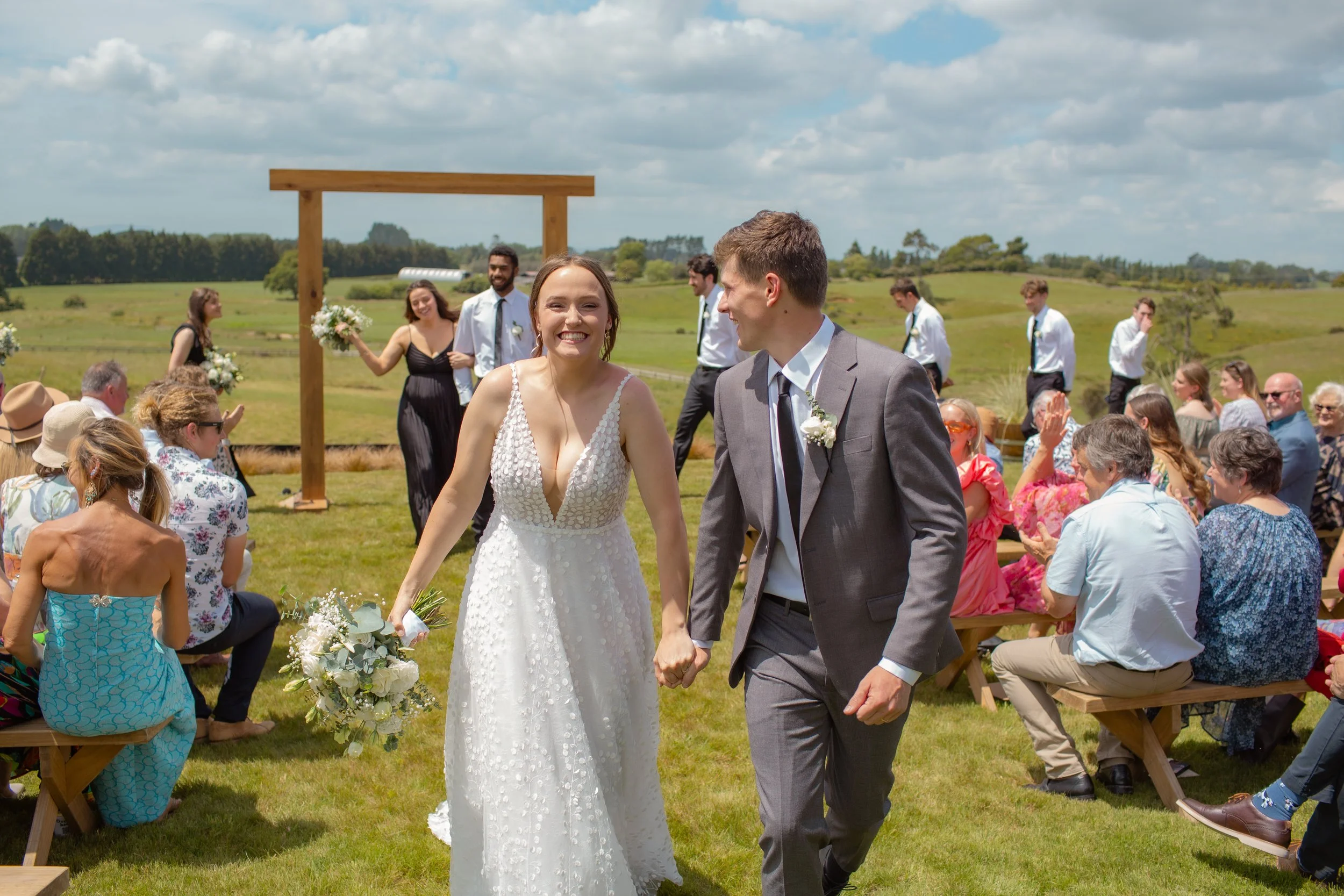 A smiling bride and groom holding hands and walking down the aisle during an outdoor wedding ceremony in a grassy field on a sunny day. The bride is wearing a white dress and holding a bouquet, while the groom is in a gray suit with a white shirt and