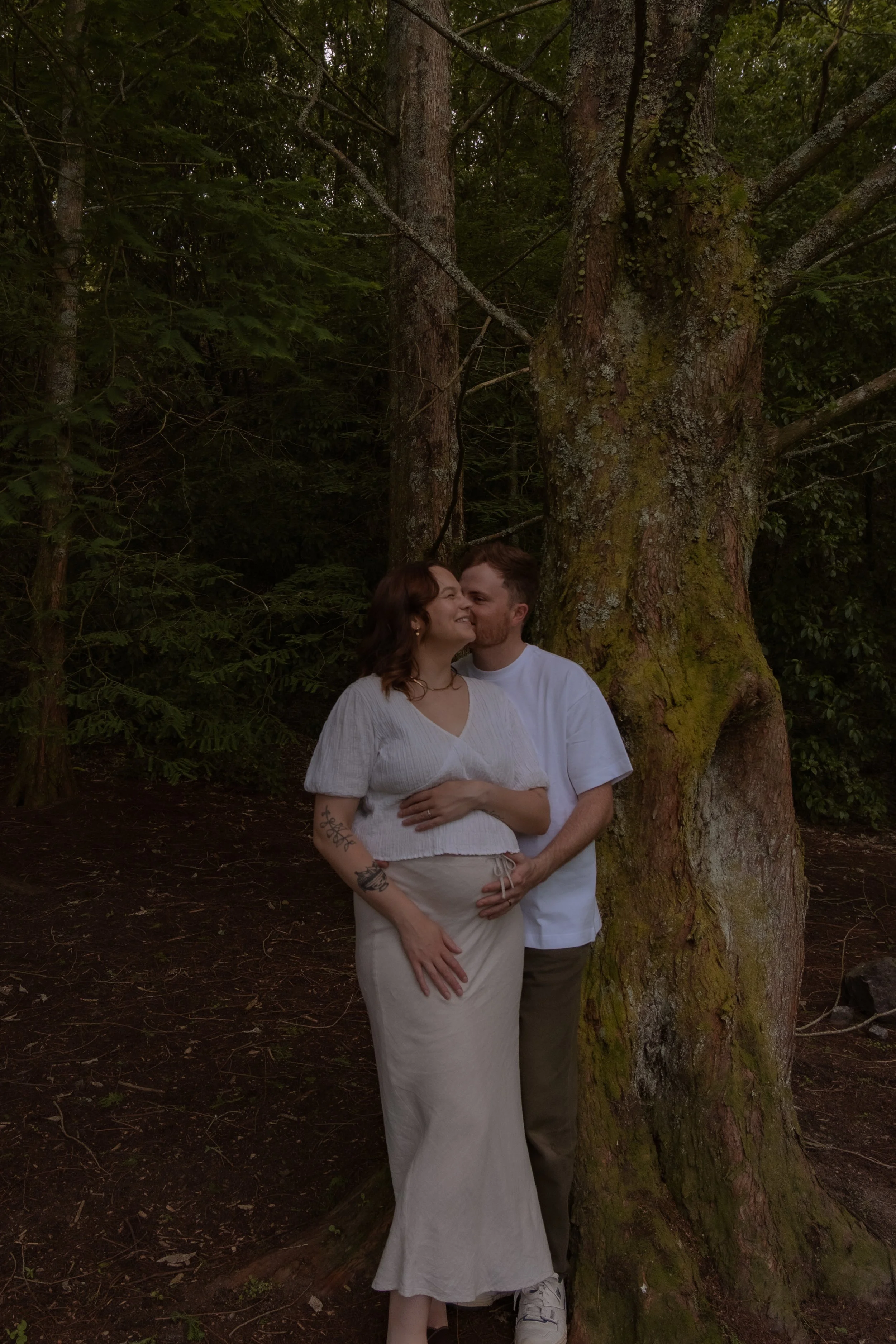 A couple embracing outdoors next to a large tree in a forest setting, with the man kissing the woman's cheek. The woman has shoulder-length brown hair, tattoos on her arm, and is wearing a white top and beige skirt. The man has short brown hair and i